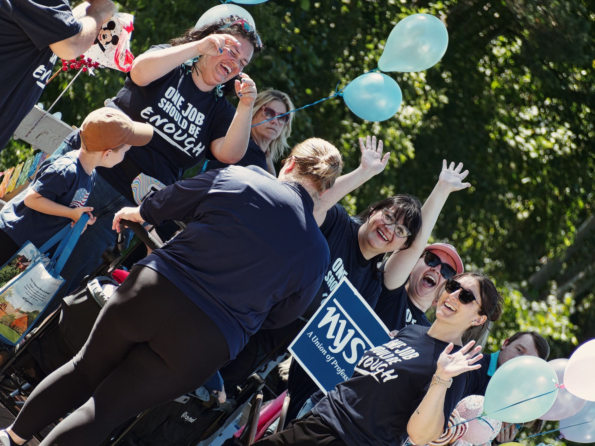 Group of people in matching navy T-shirts reading "ONE JOB SHOULD BE ENOUGH" laughing and waving during what appears to be a parade or rally, with light blue balloons floating to the right and a partially visible sign reading "NYS — A Union of Profes[sionals]" in the foreground, set against a dense green tree canopy in bright sunlight.
