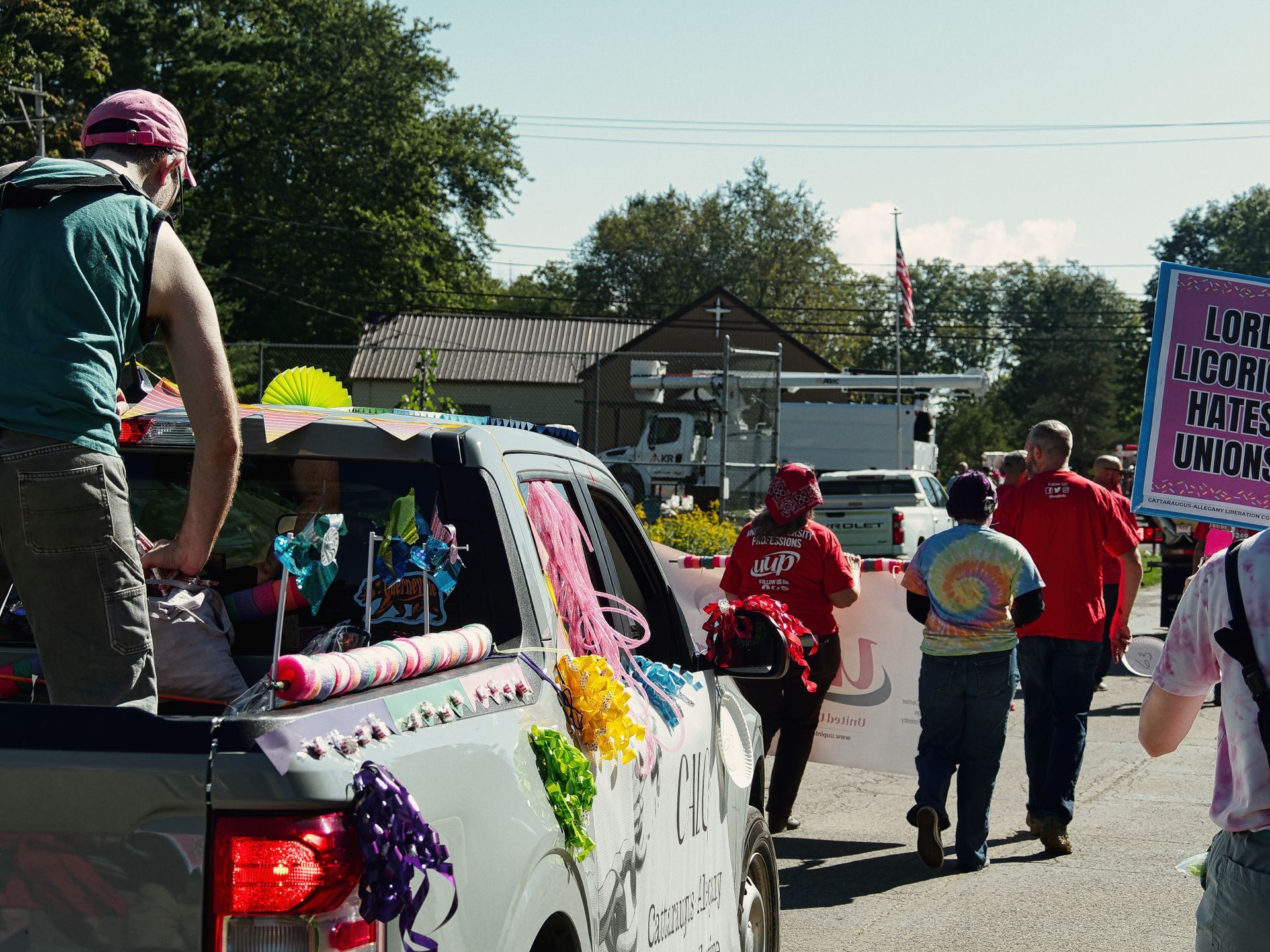 A small-town parade in progress, with a white pickup truck decorated in colorful ribbons, bows, and streamers moving down a sunny street — a man in a pink cap and teal shirt stands in the truck bed. Marchers ahead wear red shirts and a tie-dye tee, carrying a banner, while a bright pink sign on the right reads "LORD LICORICE HATES UNIONS" in bold purple letters. Trees, a barn-style building, and an American flag are visible in the background.