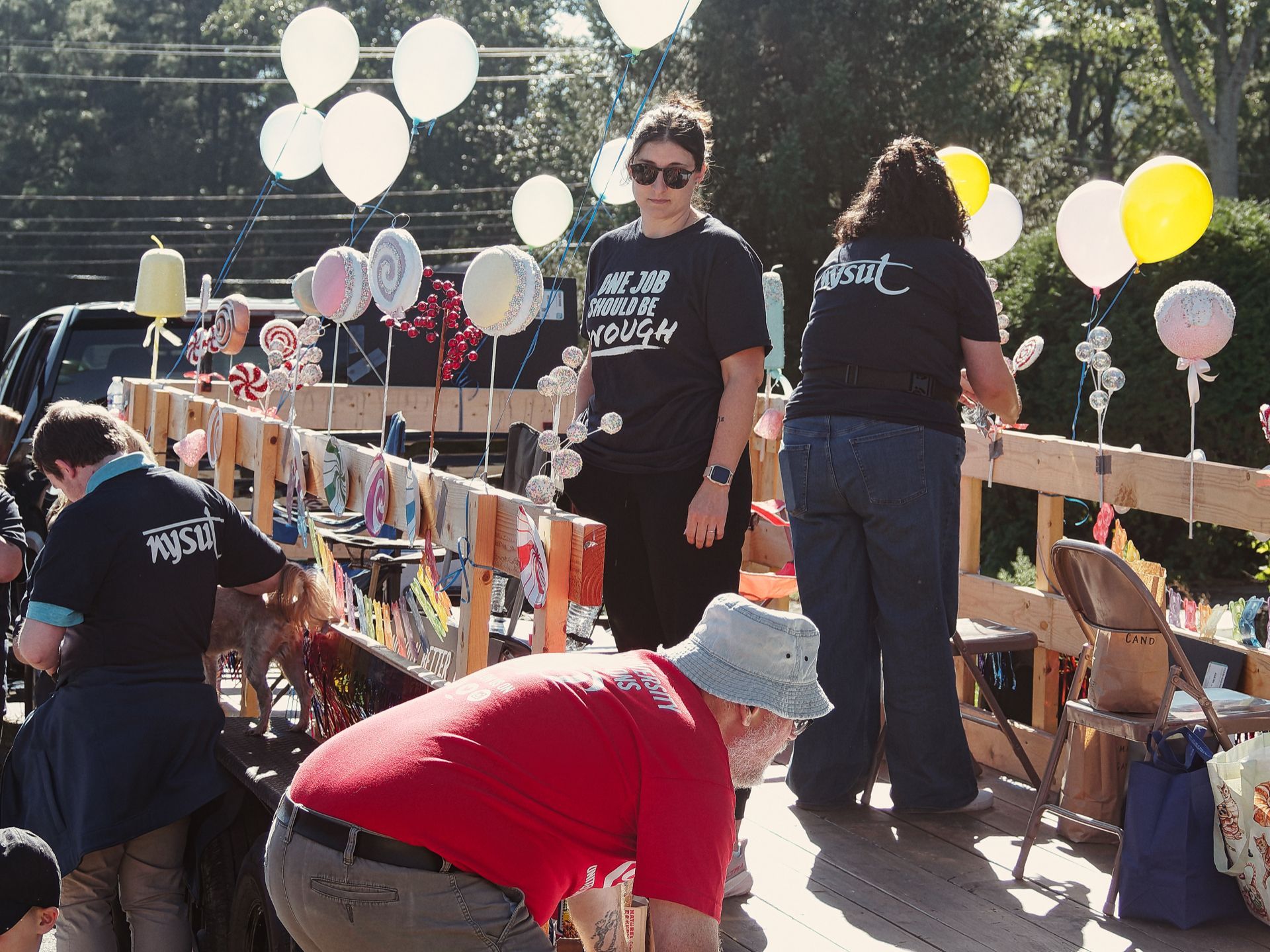 Outdoor event on a wooden platform lined with decorative oversized lollipop props and white, yellow, and pastel balloons floating overhead. Several people in black "NYSUT" shirts work the booth, including one in sunglasses whose shirt reads "ONE JOB SHOULD BE ENOUGH," while a person in a red shirt and bucket hat bends forward in the foreground. Dense trees and a parked dark truck are visible in the background.