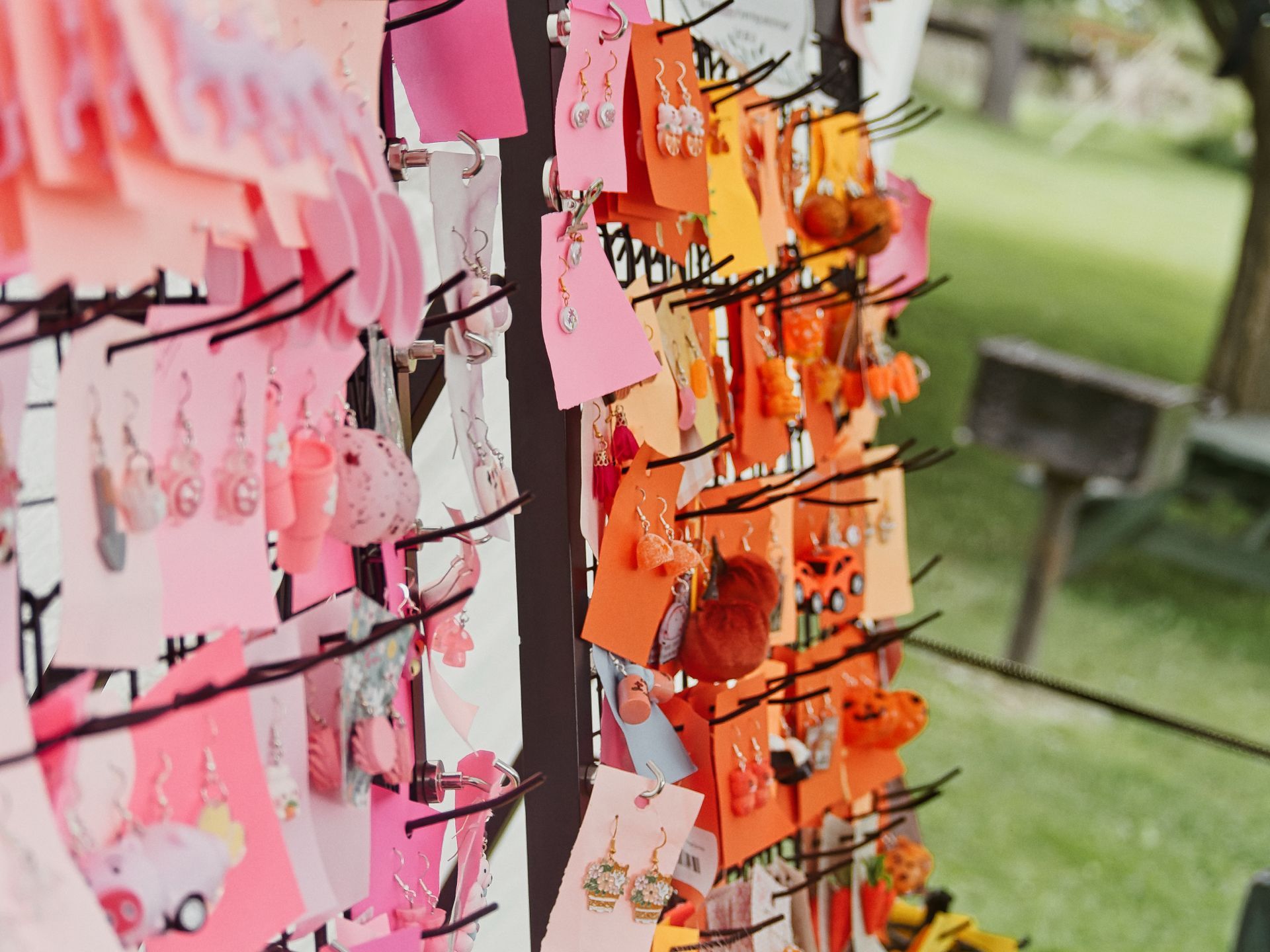 Rows of novelty earrings displayed on small pink, orange, and yellow card backings, hooked onto a rotating black metal rack at an outdoor market, transitioning from soft pinks on the left to deep oranges on the right. Green grass and a wooden bench sit blurred in the shallow depth of the background.