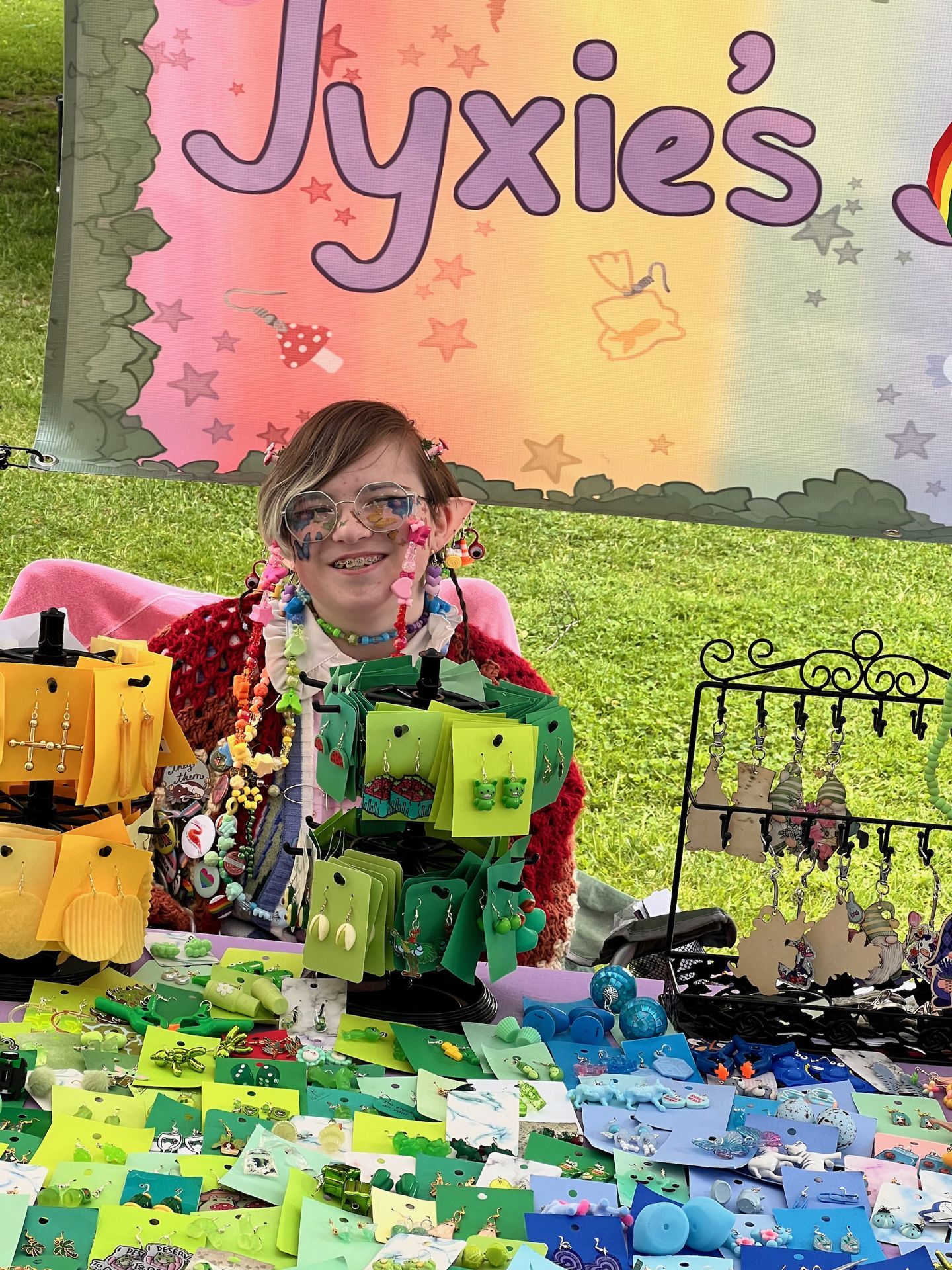 A young vendor smiling broadly behind a craft fair table overflowing with handmade earrings displayed on green, yellow, and blue card stock, with a black wire jewelry stand to the right. They wear layered colorful bead necklaces, dangly earrings, tinted round glasses, a red crochet cardigan, and face gems, holding up a green display stand of earrings toward the camera. A pastel rainbow banner reading "Jyxie's" with illustrated mushrooms and stars hangs behind them above a grass backdrop.