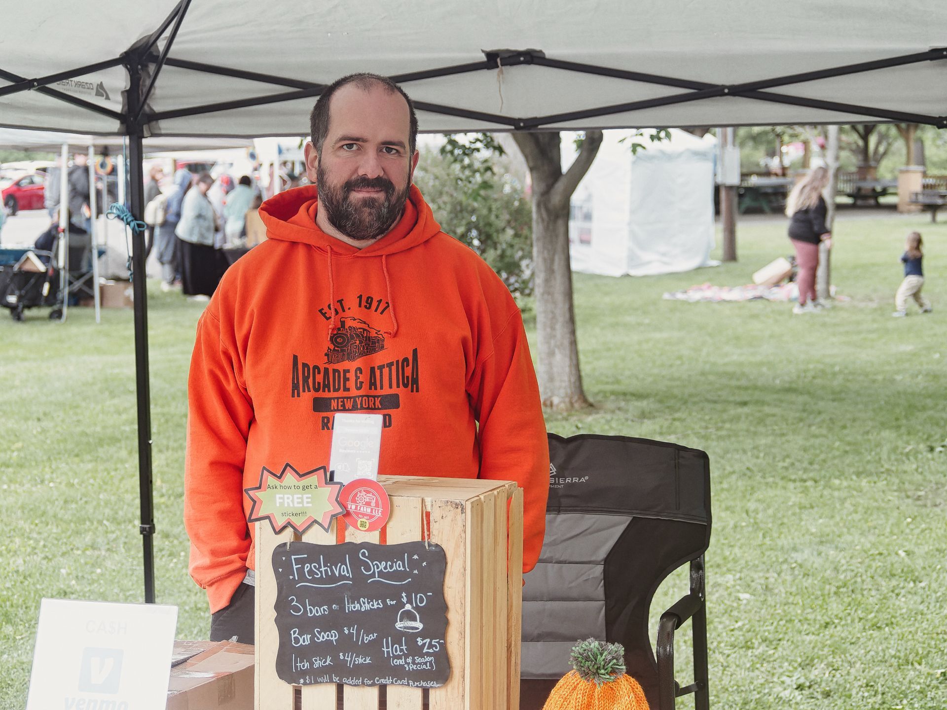 Bearded man in an orange "Arcade & Attica New York Railroad Est. 1917" hoodie standing behind a vendor table at an outdoor festival beneath a pop-up canopy. A chalkboard sign in the foreground reads "Festival Special! 3 bars or ItchSticks for $10 — Bar Soap $4/bar, Itch Stick $4/stick, Hat $2.50 (end of season special!) — a $1 will be added for Credit Card purchases," with a wooden crate display and a knitted orange pumpkin hat visible on the table; a grassy park with other attendees and tents blurs softly in the background.
