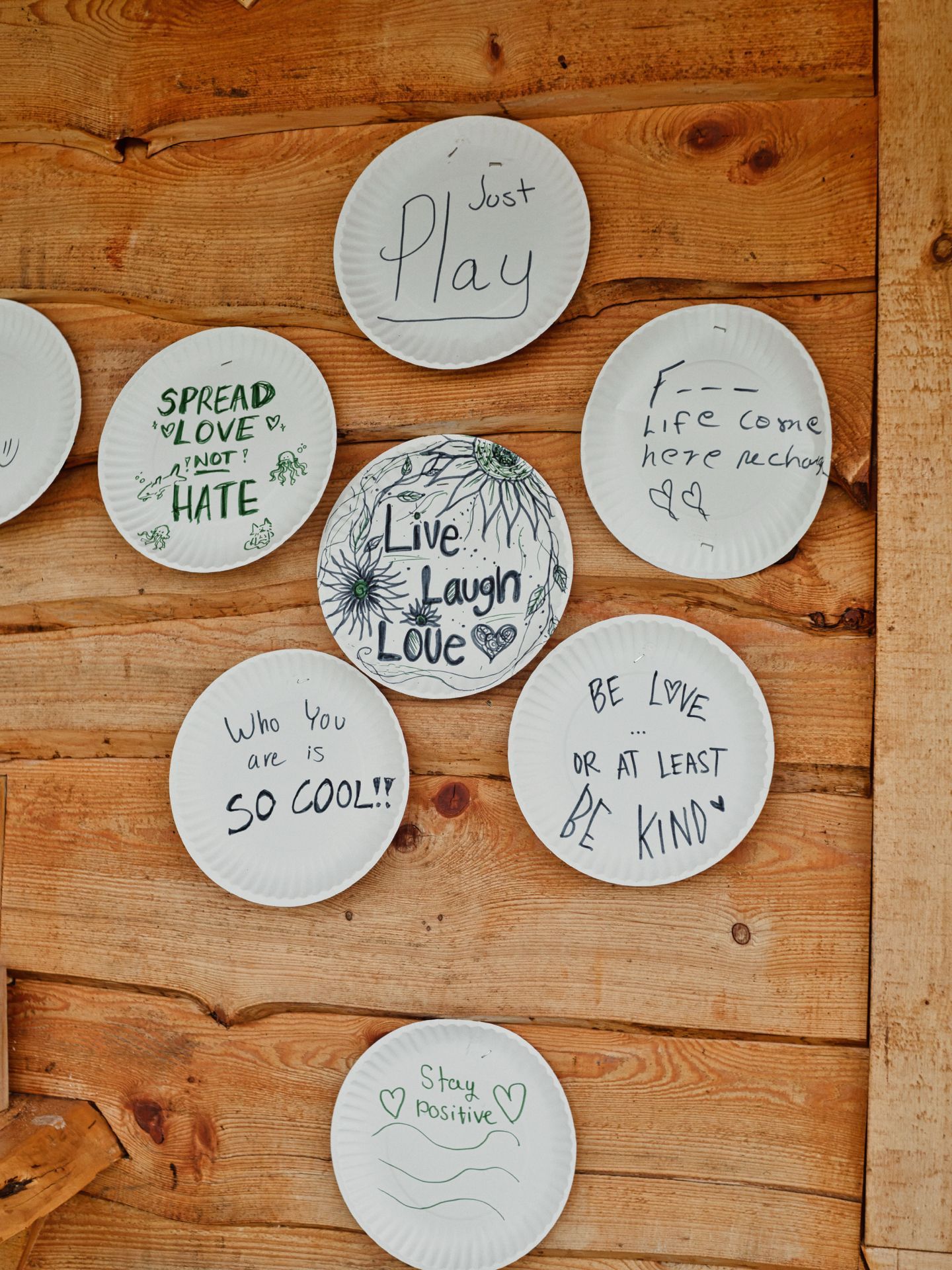 Seven white paper plates mounted on a warm pine wood wall, each hand-lettered with a phrase: "Just Play" at the top, "SPREAD LOVE ♡ NOT ♡ HATE" with small green doodles to the left, "F--- life come here recharge ♡♡" partially legible at the right, "Live Laugh Love ♡" surrounded by hand-drawn sunflowers in the center, "Who you are is SO COOL!!" at the lower left, "BE LOVE ... OR AT LEAST BE KIND ♥" at the lower right, and "♡ Stay positive ♡" at the bottom.