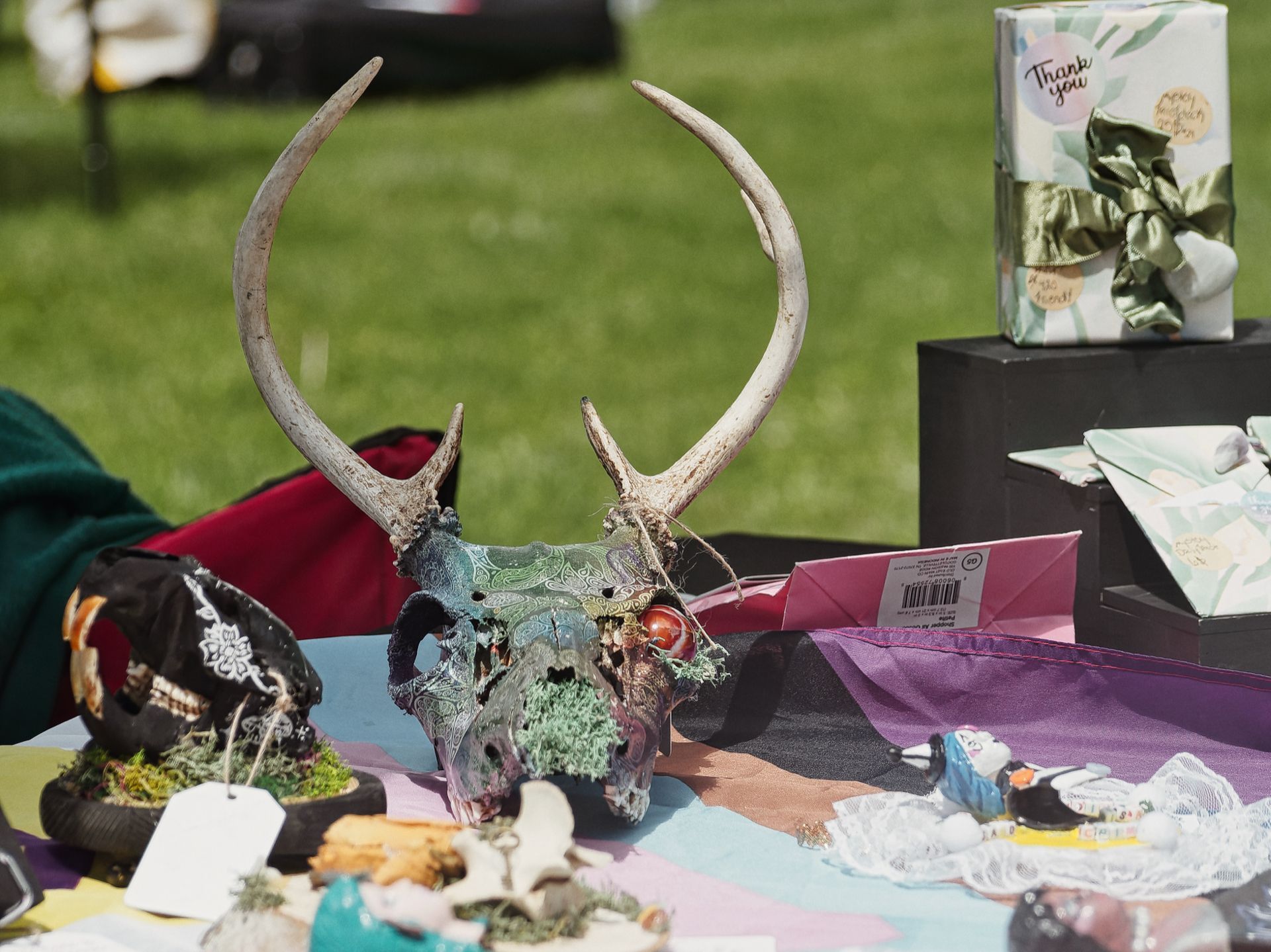 An ornately painted deer skull with full antlers — its cranium covered in teal and dark swirling patterns with moss and small red beads — sits as the centerpiece of an outdoor vendor table. Surrounding it are small figurines, tagged curiosities, fabric scraps, and a gift-wrapped box tied with a green satin bow in the upper right bearing the text "Thank you," with a sunlit grass lawn blurred in the background.