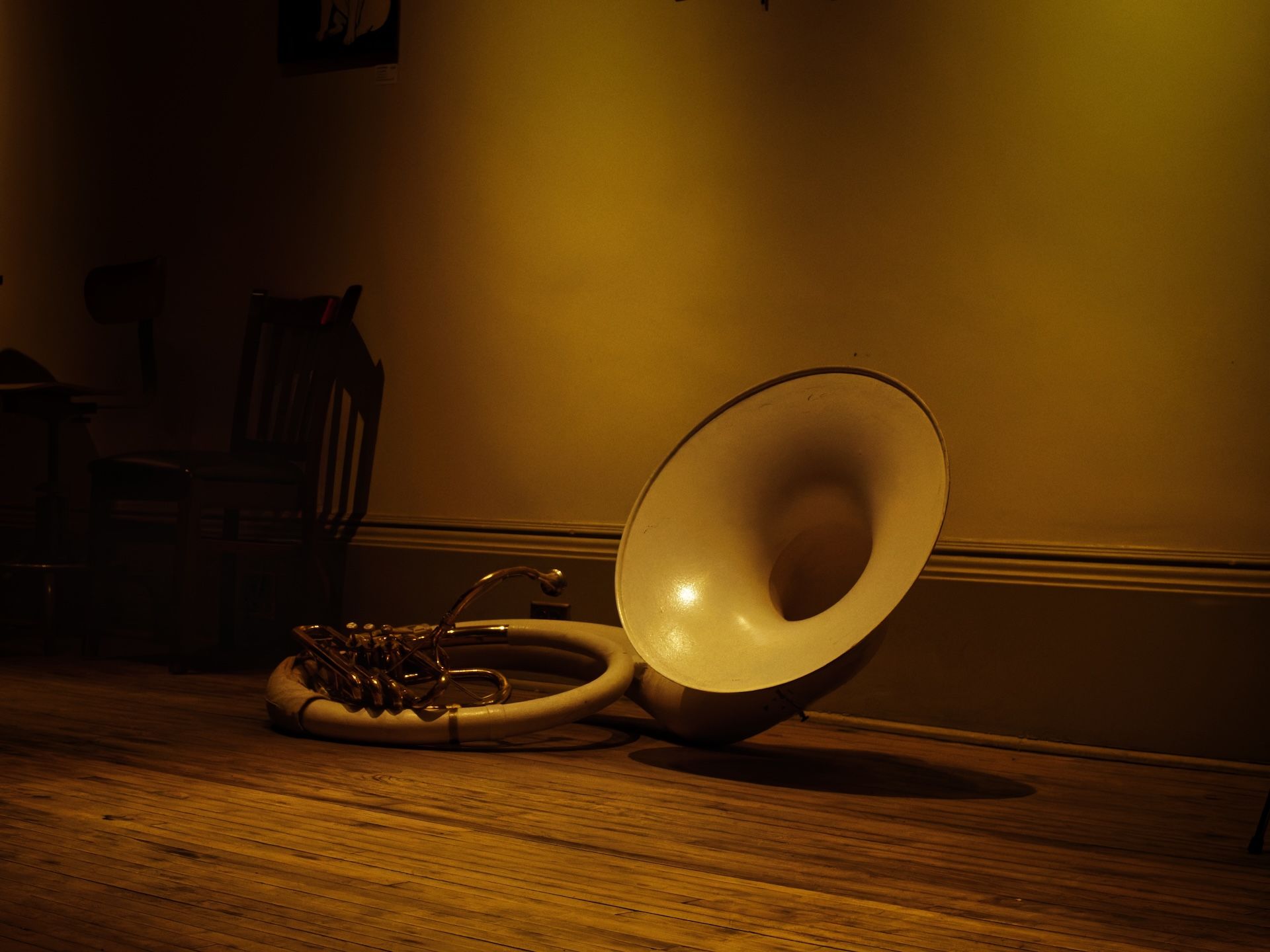 A white fiberglass sousaphone resting on its side on a worn hardwood floor, its large bell facing the camera and catching warm amber light against a yellow-washed wall. The brass valves and mouthpiece receiver curl out from the circular body to the left, with a dark wooden chair visible in the shadowy background.