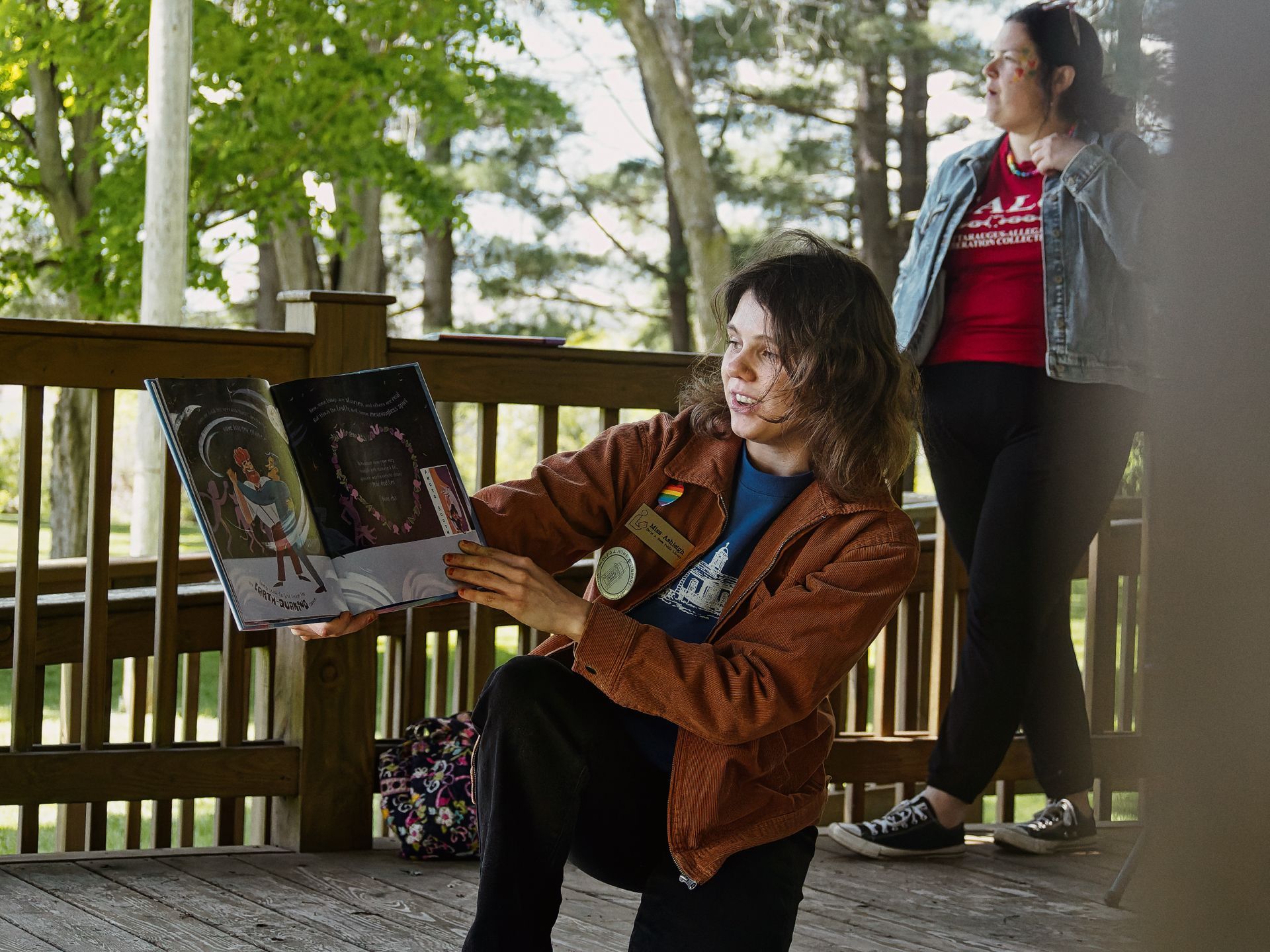 Person in a rust-orange corduroy jacket with a rainbow pin and name badge crouching on a wooden deck, holding open an illustrated children's book and reading aloud with a slight smile. Behind her, a second person in a red graphic tee and denim jacket leans against the railing, surrounded by tall trees in soft focus.