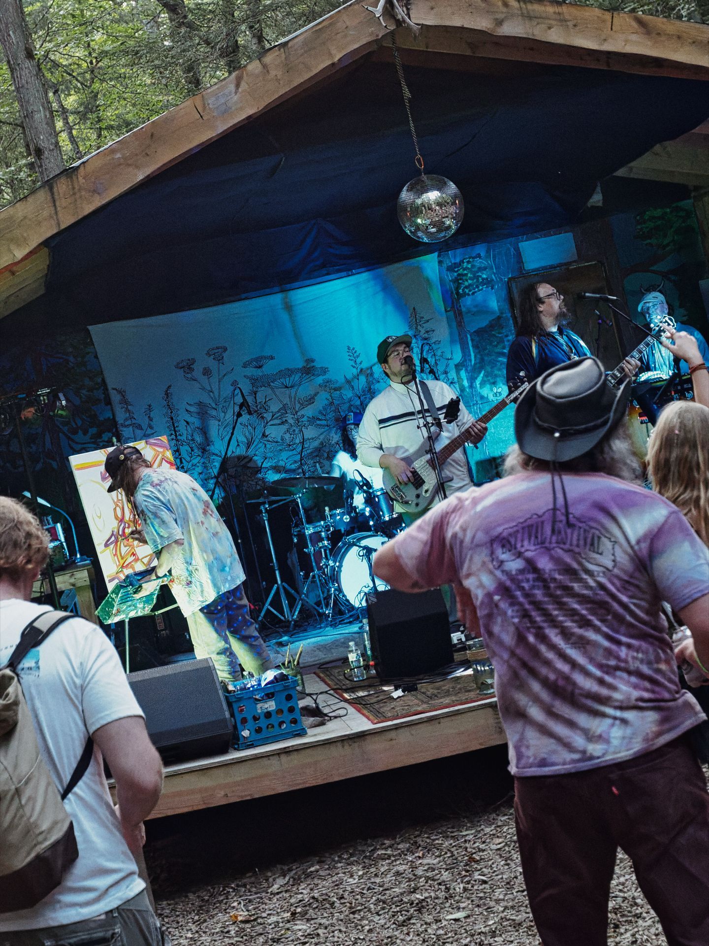 Small outdoor forest stage with a wooden roof and a mirrored disco ball hanging from the center, bathed in blue-green light. A guitarist in a white jersey and a long-haired guitarist on the right flank a drum kit at the back, while a paint-splattered performer works on a colorful canvas at stage left. In the foreground, two audience members watch — one in a tie-dye shirt reading "FESTIVAL" with a wide-brim hat, the other carrying a backpack — with tall trees visible beyond the stage canopy.