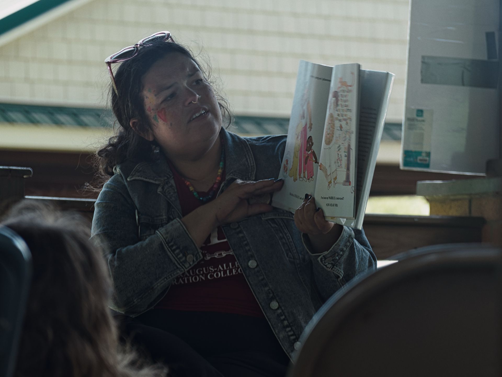 Woman with sunglasses pushed up on her head and colorful face paint holds an open picture book outward toward an audience, mid-reading aloud. She wears a denim jacket over a red shirt partially reading "...AUGUS-ALL... RATION COLLE..." and a beaded necklace; the book's illustrated pages show a dark-skinned girl figure with text visible including "Do I have my MARVELOUS adventure?" and "NOW WE DO THIS." Two listeners are partially visible in the foreground, blurred in the shallow depth of field.