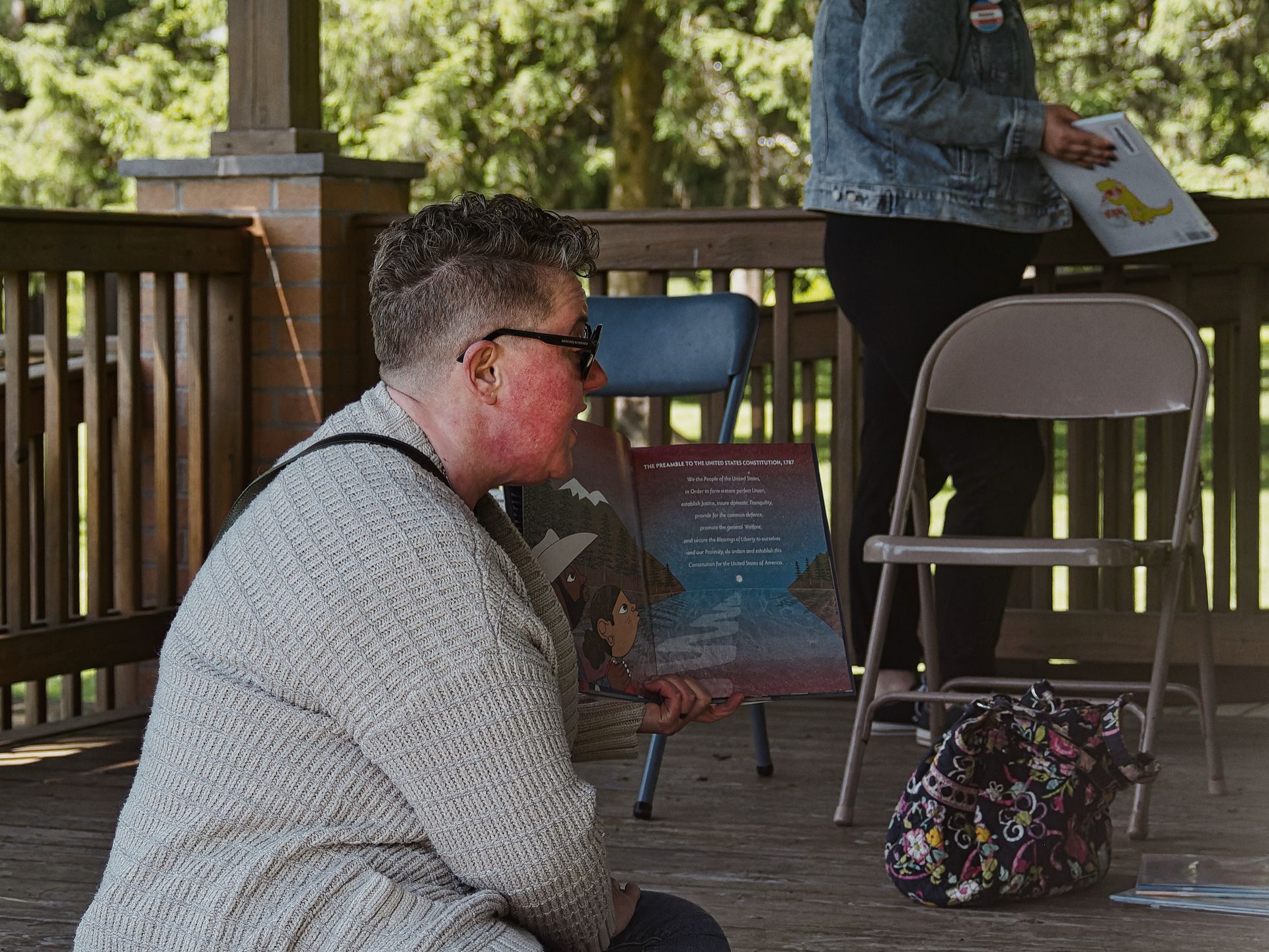 Person in a gray knit sweater and black glasses, seated on a wooden deck, holding open a children's book to a page headed "THE PREAMBLE TO THE UNITED STATES CONSTITUTION, 1787," with illustrated night-sky artwork visible on the left page. Behind them, a second person in a denim jacket holds a book featuring a yellow dinosaur illustration, partially cropped at the upper right. Two folding chairs and a floral Vera Bradley bag sit on the deck, with a wooden railing and lush green trees filling the background.