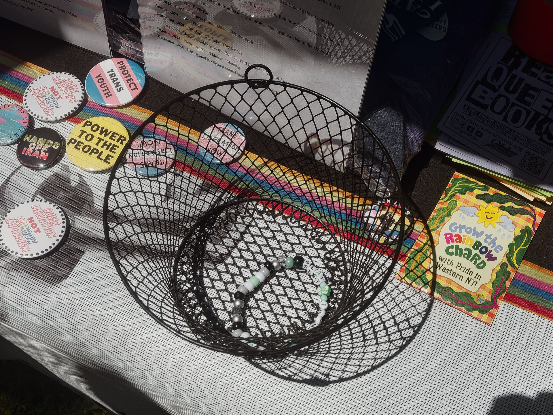 An outdoor table spread with activist and pride materials: a black wire basket holds beaded bracelets and buttons reading "Trans Liberation Now" and "Nothing About Us Without Us," while loose buttons to the left read "Protect Trans Youth," "Power to the People," and "Hands Off Iran." A seed packet to the right reads "Grow the Rainbow Chard with Pride in Western NY!" in colorful lettering, and a rainbow-striped cloth runs beneath everything.