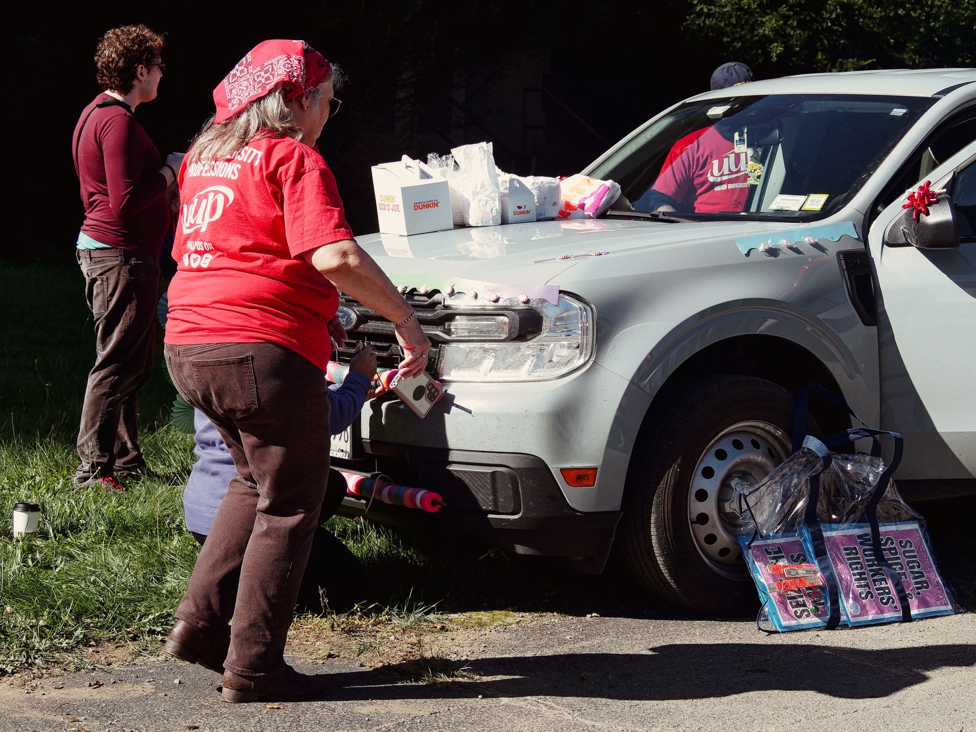 Woman in a red "University Professions" t-shirt and red bandana leans over the hood of a white SUV loaded with Dunkin' bags and cups, while a second person in dark red stands to the left in conversation. On the ground by the front tire, three tote bags read "Sugar Splices," "Sugar Workers Rights," and "Sugar."