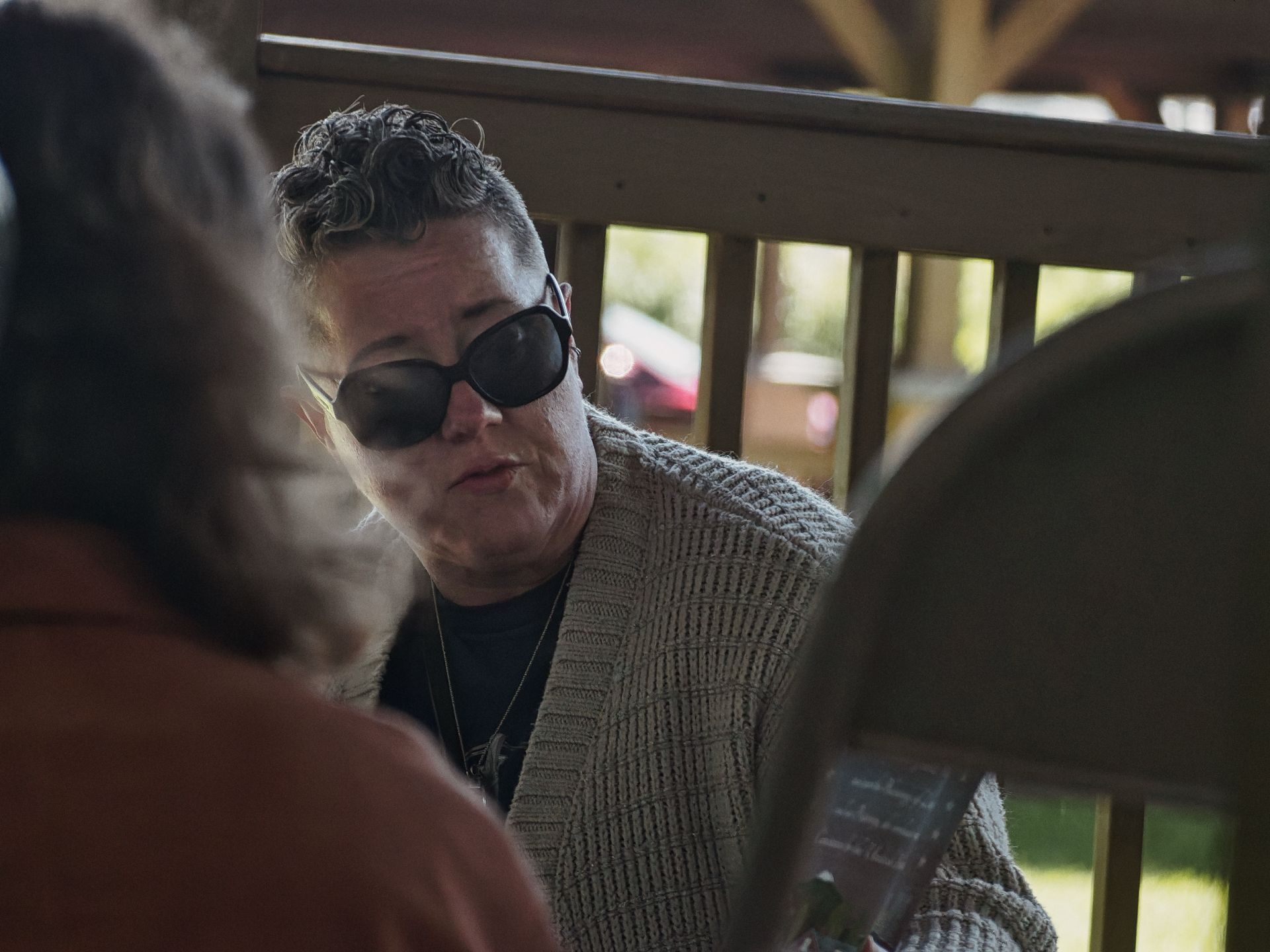 Person with short curly hair, dark oversized sunglasses, and a chunky knit cardigan over a black top, looking downward in conversation with others whose backs and shoulders blur the foreground. A wooden deck railing and soft green foliage fill the background.