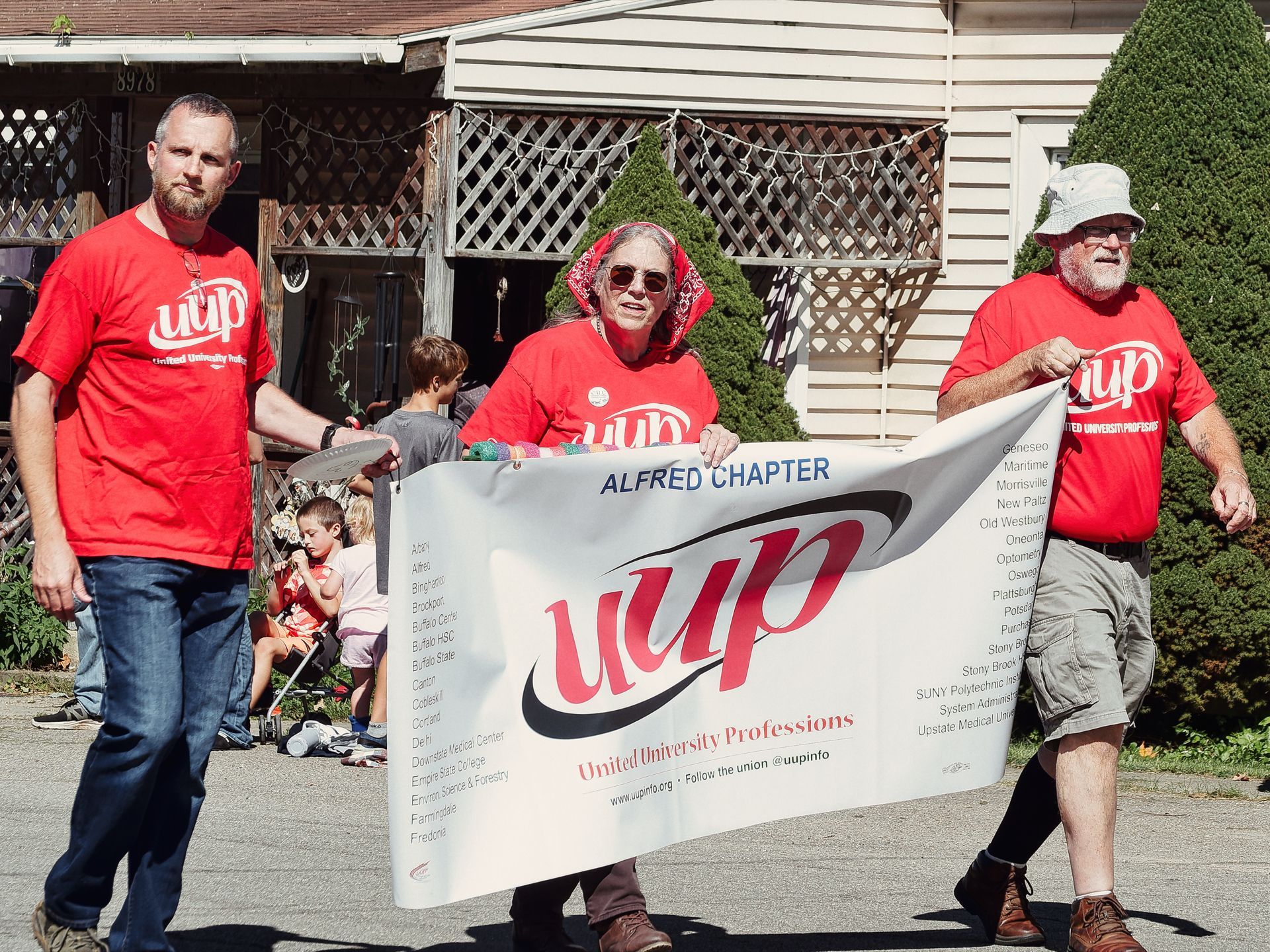 Three people in matching red UUP (United University Professions) shirts march in a parade, holding a large white banner reading "Alfred Chapter — UUP United University Professions" with a list of chapter locations along both edges. A beige house numbered 8978 and dense evergreen shrubs fill the background, where children sit watching from the sidewalk.