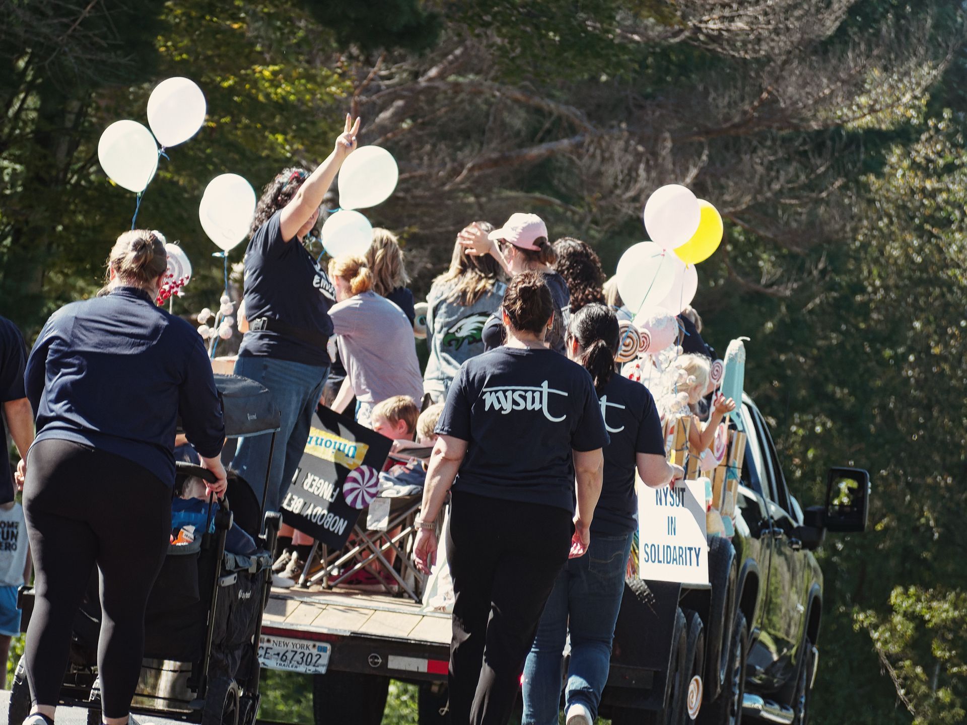 Parade float on a New York-plated trailer, pulled by a dark SUV, with a sign reading "NYSUT IN SOLIDARITY" on the right side and a reversed banner reading "NOBODY DARES" on the float's back edge, decorated with white and yellow balloons and candy-themed props. Several people in black "NYSUT" t-shirts walk alongside and ride the float, one raising a peace sign, while a person pushing a stroller walks at the far left, all moving away from the camera down a tree-lined road.