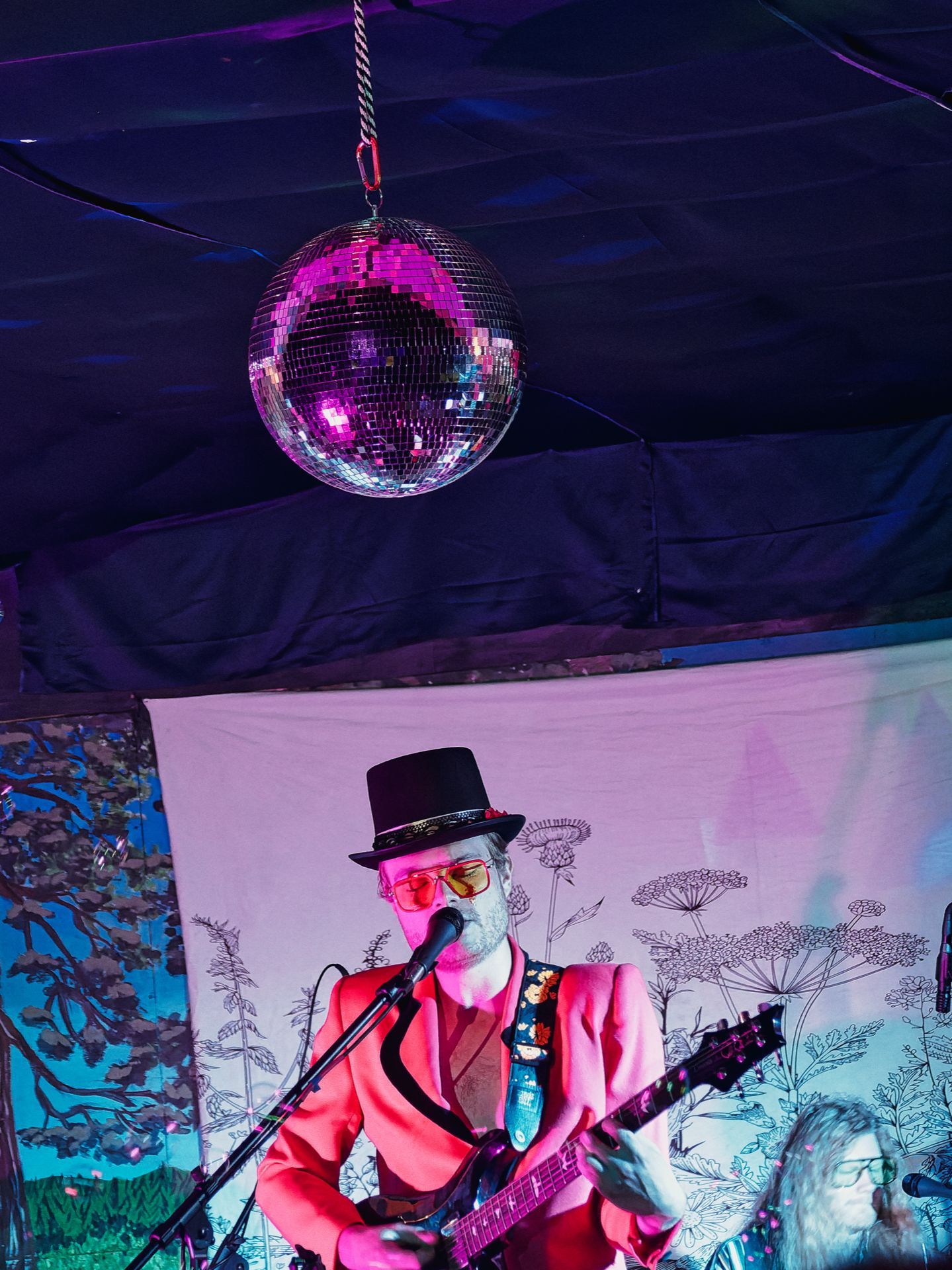 A guitarist in a hot pink blazer, black top hat, and red-tinted round glasses plays into a microphone beneath a mirror ball casting magenta light from above, with a illustrated botanical-print backdrop behind him and a second performer partially visible in the lower right corner.