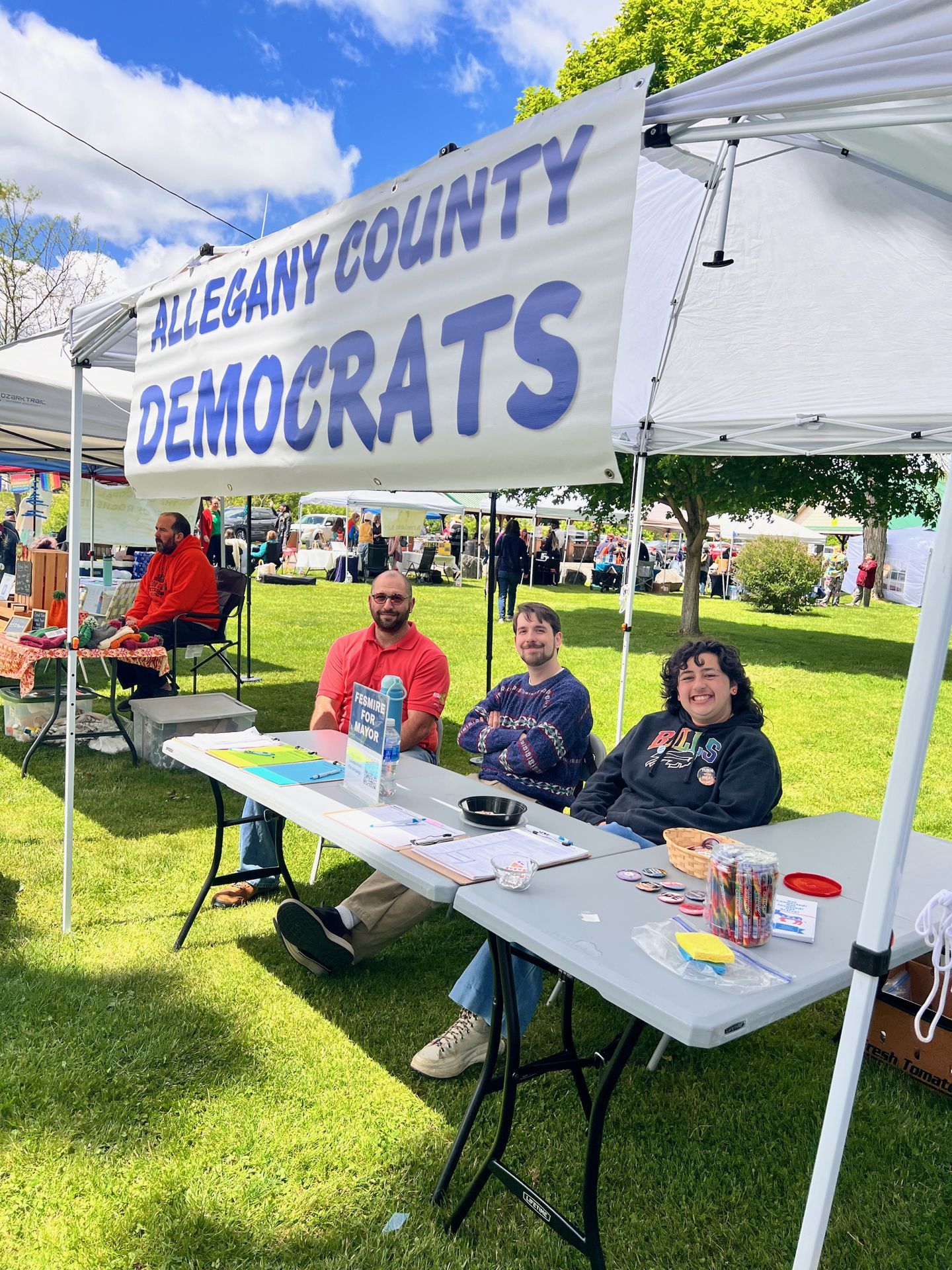 Three people seated at a folding table beneath a white canopy tent at an outdoor fair, under a large banner reading "ALLEGANY COUNTY DEMOCRATS." The table holds clipboards, pins, markers, and a basket of buttons; a small sign reading "PERKINS FOR MAYOR" is visible at the left end. Other vendor tents and fairgoers fill the sunny, green-lawned background.