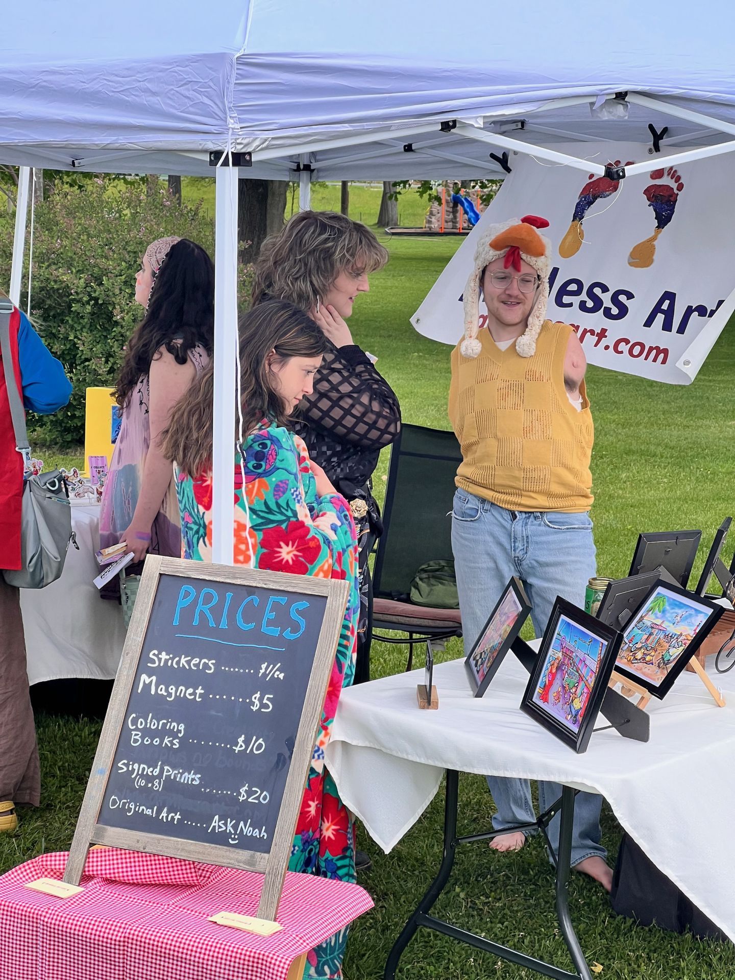 Outdoor art market booth under a white canopy, where a young vendor wearing a yellow knit vest and a chicken hat gestures animatedly toward two customers — a woman in a black mesh top and a child in a floral dress. A chalkboard sign in the foreground reads "PRICES / Stickers ......$1ea / Magnet ......$5 / Coloring Books ......$10 / Signed Prints (10x8) ......$20 / Original Art ..... Ask Noah." Framed colorful prints are displayed on a white-draped table to the right, and a partially visible banner behind the vendor reads "...less Art... ...art.com."