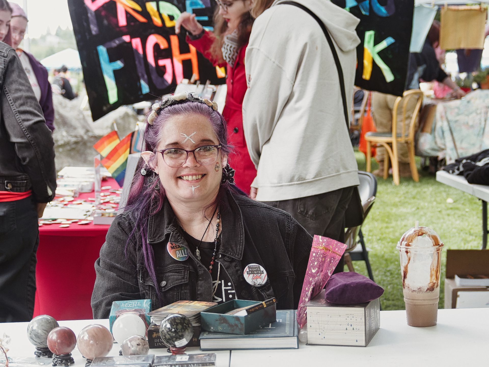 Woman with purple hair, small decorative horns, pointed ear accessories, and white cross face paint smiles from behind a vendor table at an outdoor Pride event, wearing a black denim jacket with enamel pins reading "HEAL ALL" and "TRANS RIGHTS OR I BITE." The table holds crystal spheres, stacked books, a teal box, and an iced chocolate drink, with a black banner reading "PRIDE FIGHT" and a small rainbow flag visible behind her.