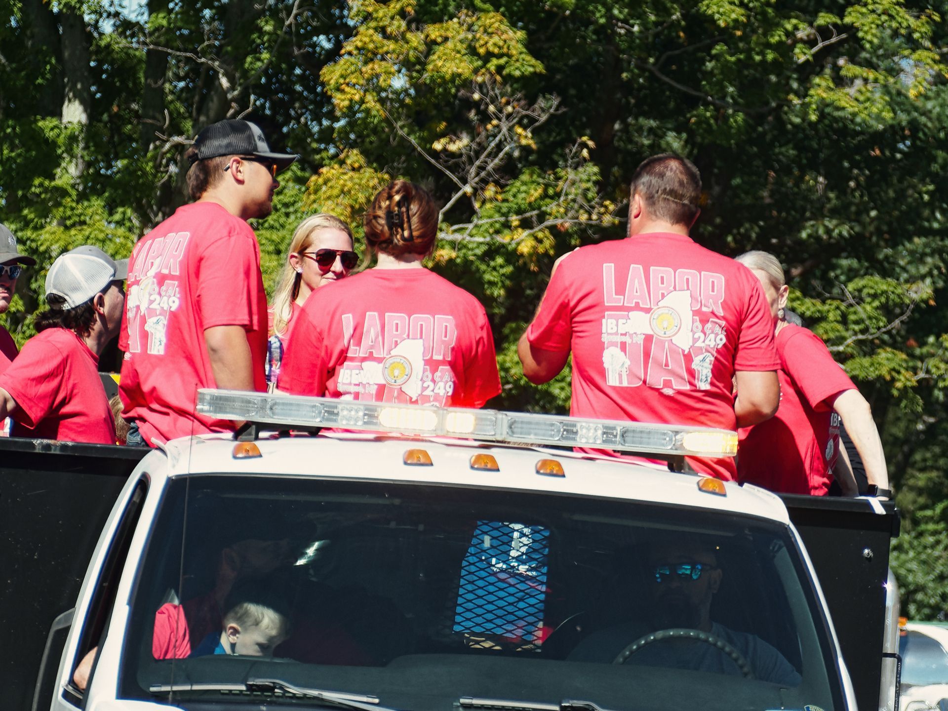 Group of people riding in the bed of a white flatbed truck, most wearing matching red "LABOR DAY IBEW 249" t-shirts, with dense green trees filling the background. A driver and passenger are visible through the windshield below, and a blonde woman in red sunglasses faces forward among the group on the truck bed.