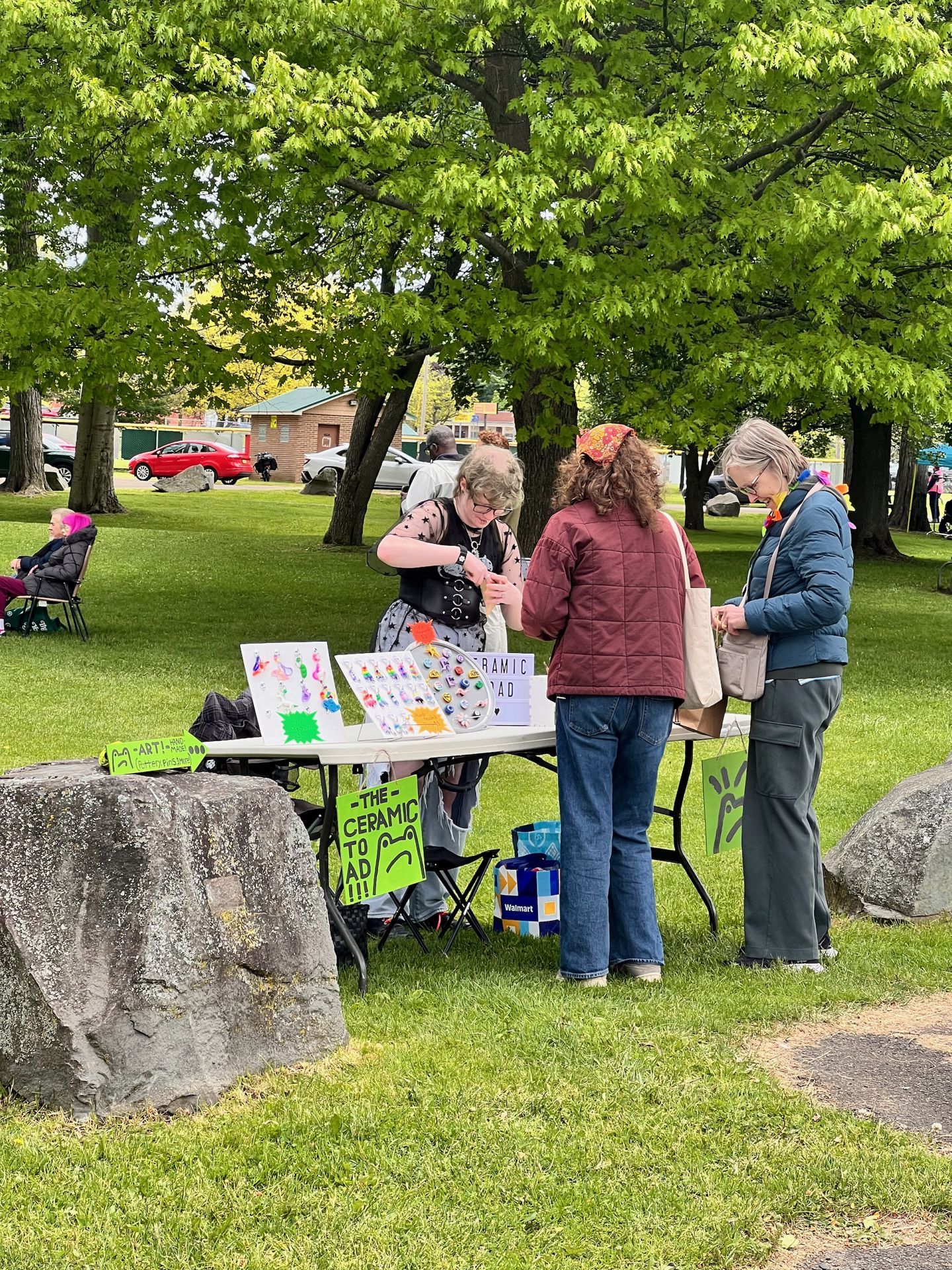 Three people gathered around a folding table at an outdoor park vendor booth, browsing colorful handmade art and ceramic pieces displayed on white boards. A neon green sign on the table reads "THE CERAMIC TOAD!!!" and a smaller sign to the left reads "=ART!="; a Walmart bag sits on the grass below, with large stone boulders framing the foreground and leafy trees filling the background.