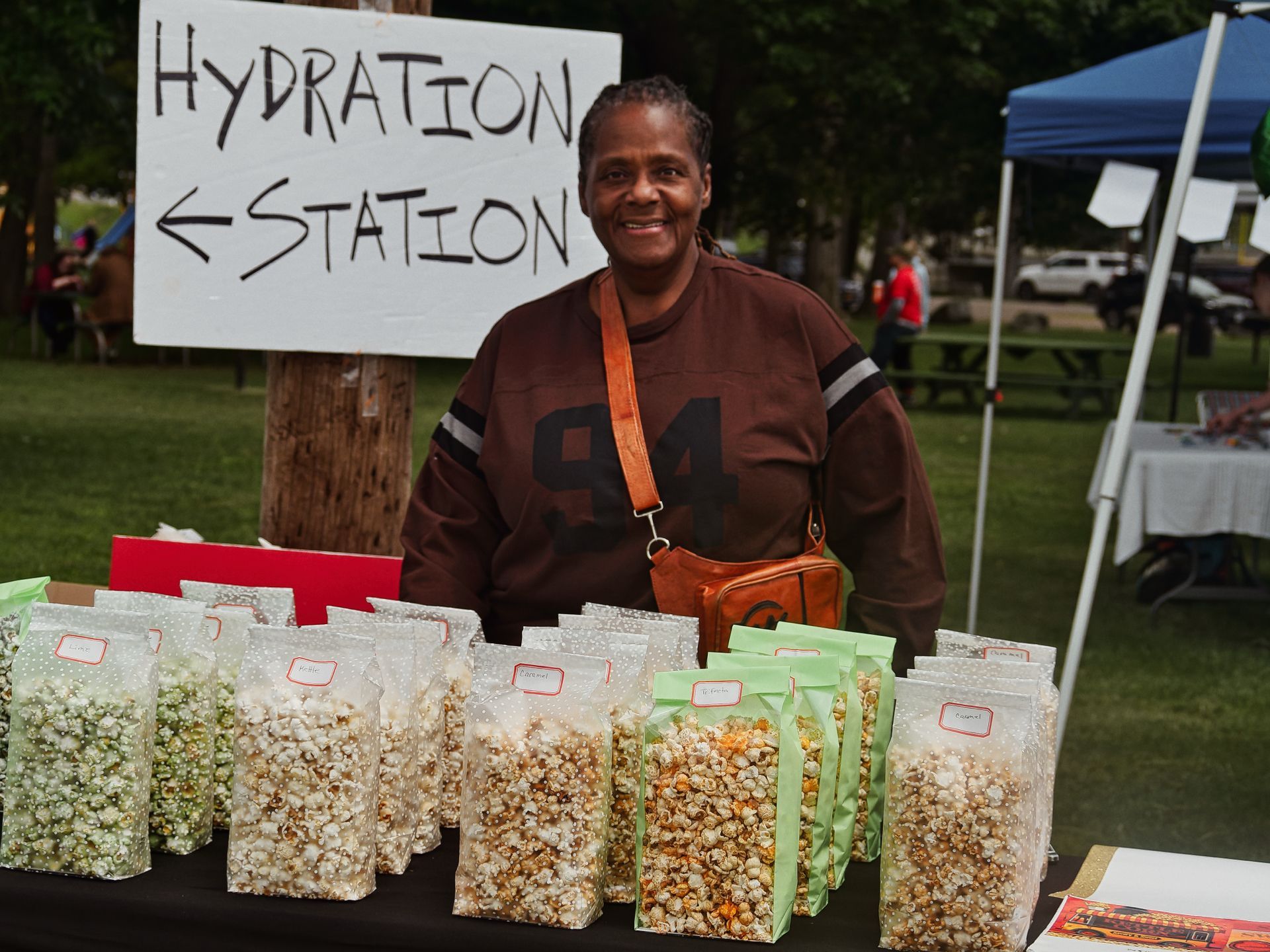 A smiling vendor in a brown #94 jersey stands behind a table lined with clear and green-topped bags of popcorn in various flavors, each with a small label, at an outdoor market. Behind her, a handwritten sign on a post reads "HYDRATION STATION ←," with green grass, trees, and a blue canopy tent blurred in the background.