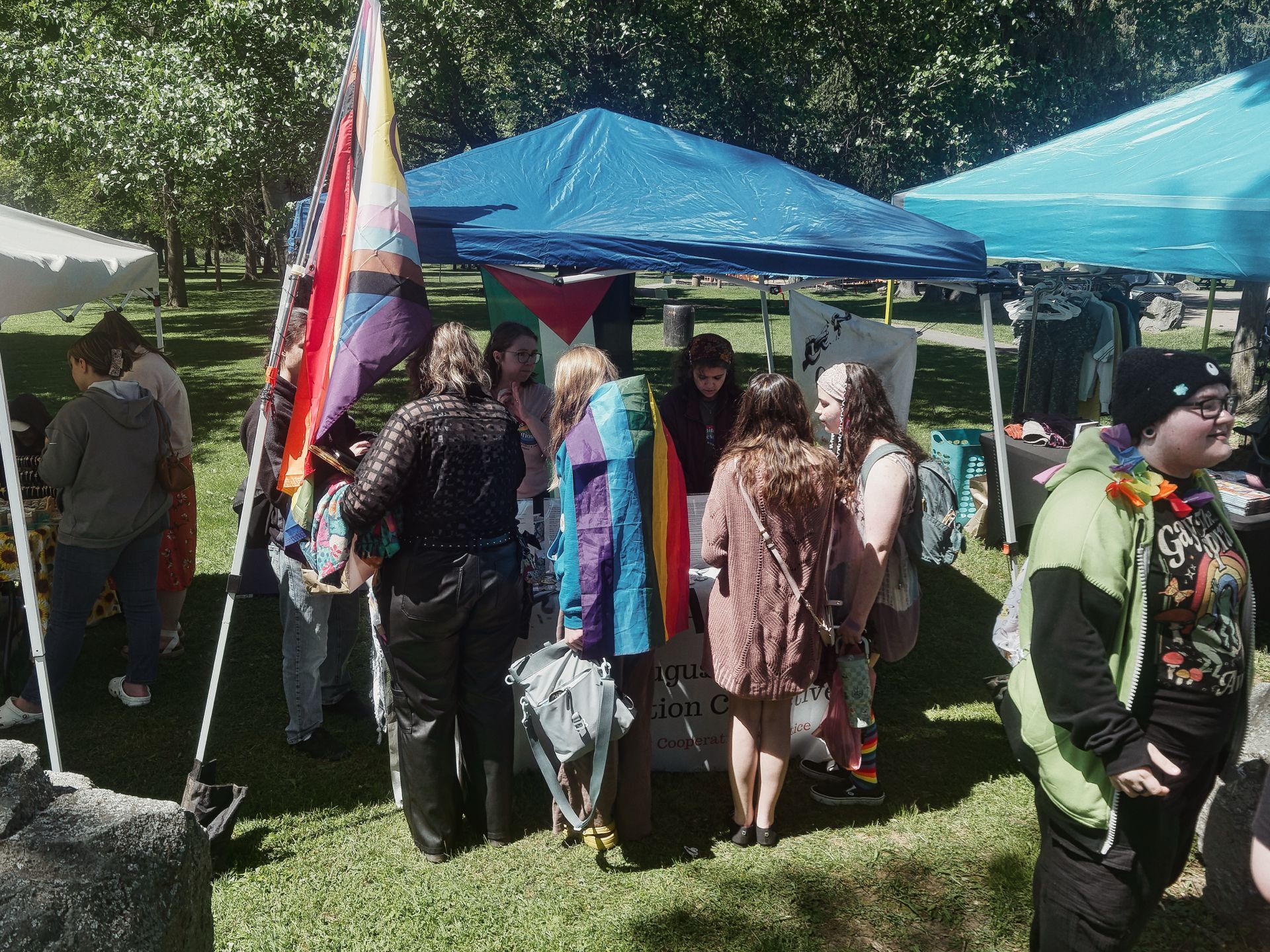 An outdoor fair or market on a sunny grassy field, with a cluster of people gathered under a blue pop-up canopy around a table bearing a partially visible sign reading "...ugu... ...tion Co...tive / Coop..." — a Pride-themed event, with a large Progress Pride flag flying at the left and a trans Pride flag draped over one attendee's shoulders. At the far right, a person in a lime-green hoodie and a rainbow clown-ruffle collar walks toward the camera, and additional vendor tents and tree canopy fill the background.