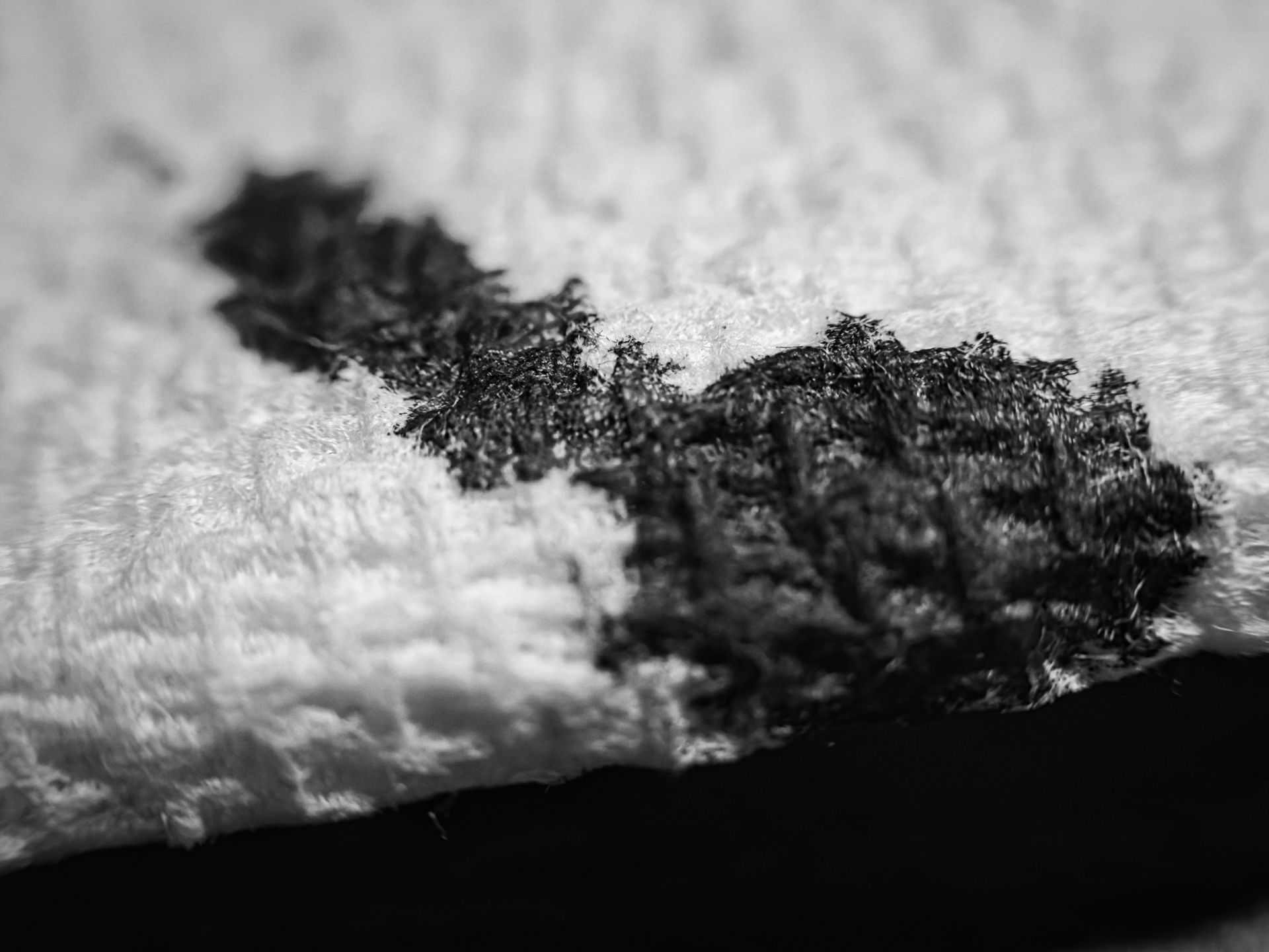 Black-and-white extreme close-up of an ink blot on a paper towel, its jagged, feathery edges looking nearly like a aerial view of a treeline, with the towel's edge and a black surface visible at the bottom of the frame.