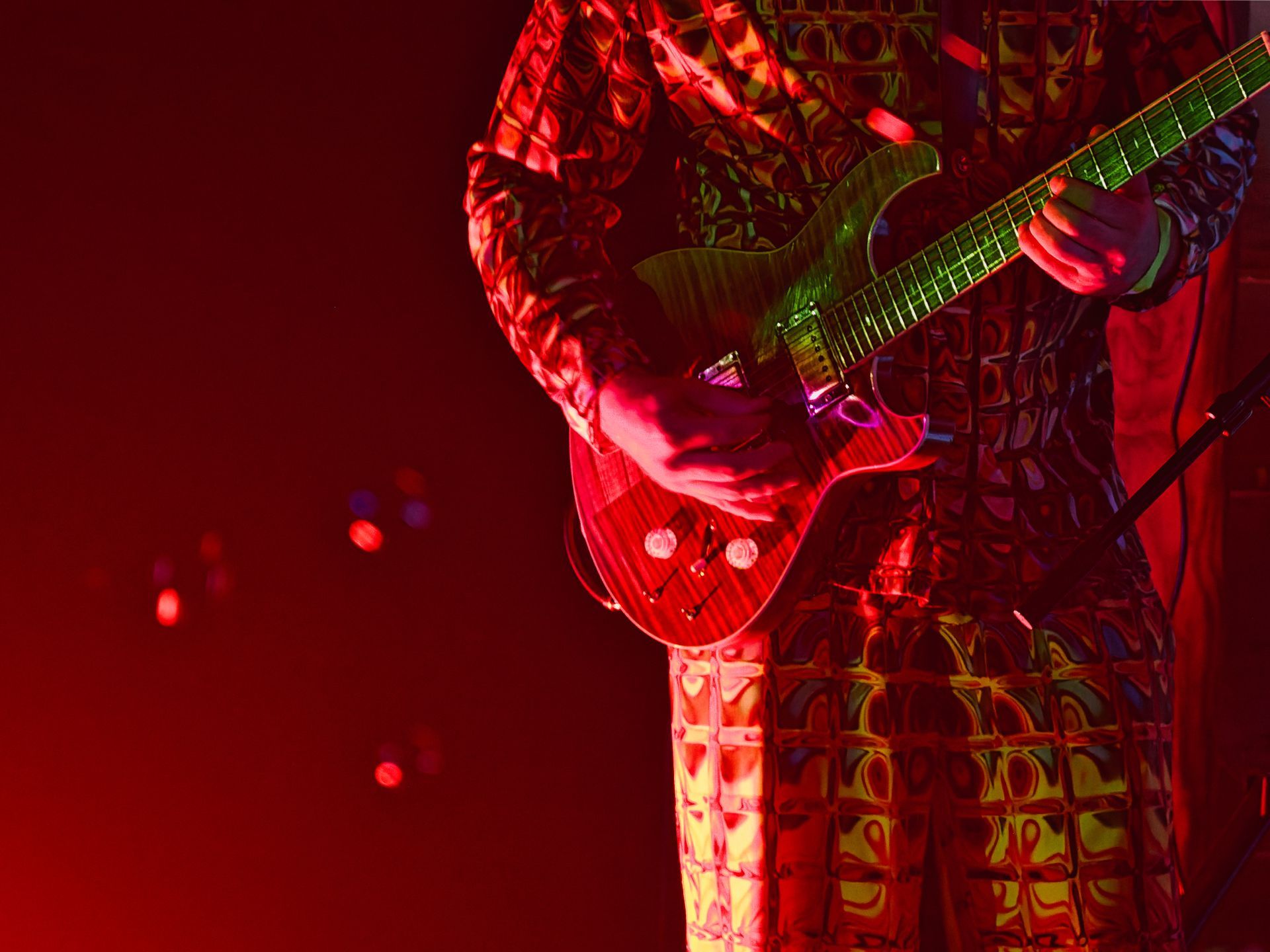 Torso and hands of a guitarist in a bold psychedelic-print shirt, playing a deep red electric guitar under saturated red stage lighting, cropped just below the chin and above the knees. Bokeh stage lights glow faintly in the deep red background to the left.