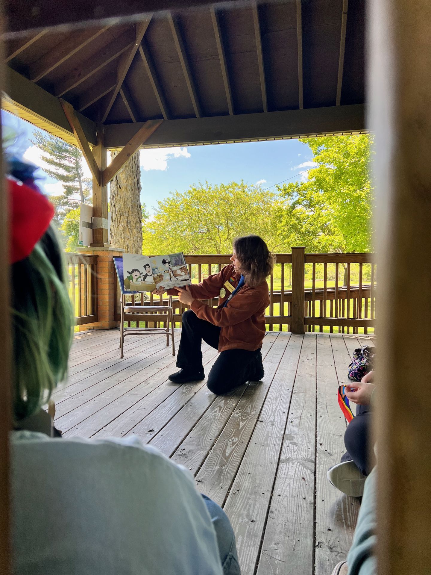 Person in a rust-brown hoodie kneeling on a wooden deck under a pavilion roof, holding open an illustrated picture book toward a small audience, shot from just inside a doorway. In the foreground left, the back of a listener wearing a red hat with green-dyed hair; to the right, another listener holds a rainbow-striped object. Bright green spring trees and a blue sky fill the open background beyond the deck railing.
