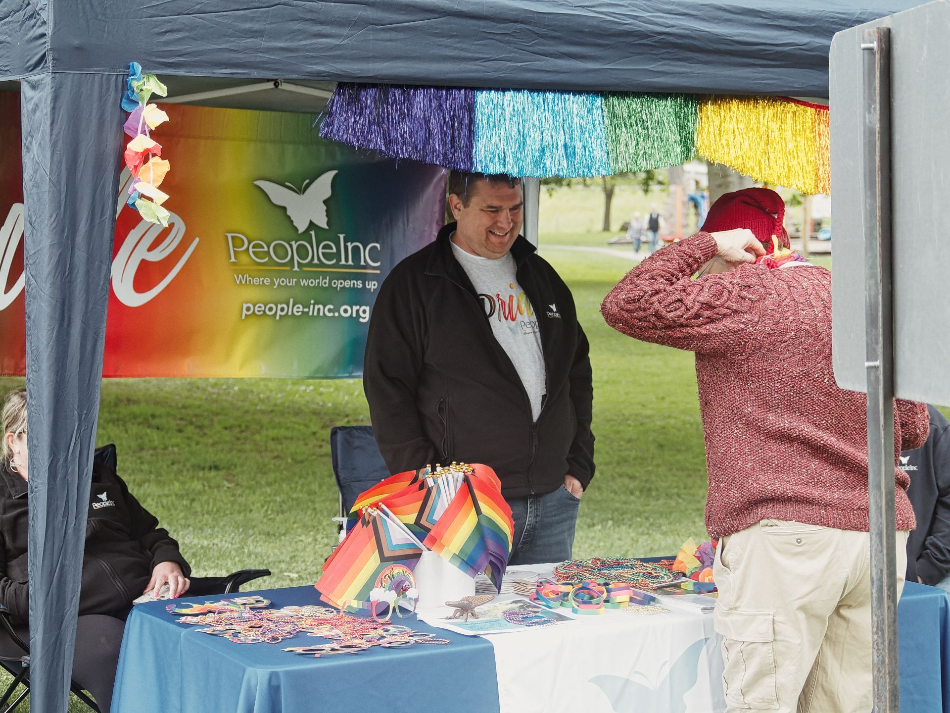 An outdoor Pride event booth under a navy canopy decorated with rainbow tinsel fringe, staffed by a smiling man in a black jacket behind a blue-draped table loaded with Pride flags, stickers, beads, and bracelets. A rainbow banner behind him reads "PeopleInc — Where your world opens up — people-inc.org," while a visitor in a red knit cap and mauve cable-knit sweater browses from the right foreground.
