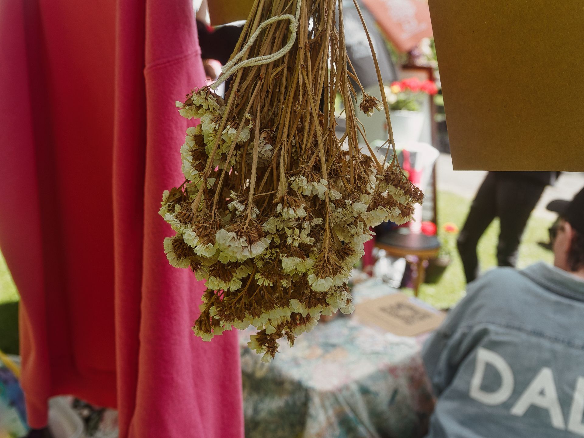 A bundle of dried white flowers hung upside down by twine, centered in the frame against a blurred market scene — a bright pink garment hangs to the left, and a person in a gray sweatshirt reading "DA" sits to the lower right, with green grass and vendor tables soft in the background.