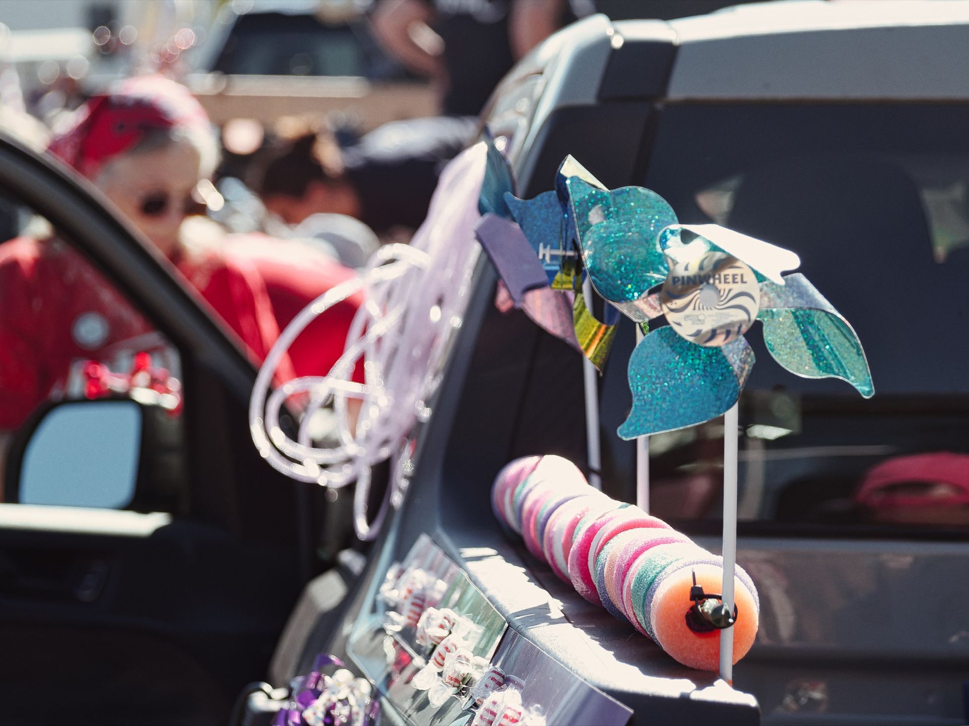 A teal glitter pinwheel labeled "PINWHEEL" mounted on a white stick sits atop a car's trunk or door, alongside a rolled pastel candy log, a white looping toy, and a tray of wrapped candies — a trunk-or-treat setup in bright afternoon sun, with a child in red and a red bandana blurred in the shallow background left.
