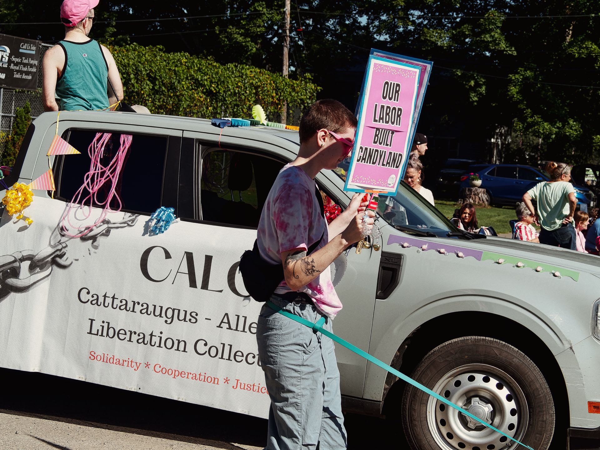 A parade participant in a tie-dye shirt holds a pink sign reading "OUR LABOR BUILT CANDYLAND" while walking alongside a decorated white vehicle bearing a banner for "CALC — Cattaraugus - Alle[gheny] Liberation Collec[tive]" with the tagline "Solidarity * Cooperation * Justic[e]." A person in a pink cap sits atop the vehicle, and onlookers line the background beneath a canopy of trees.