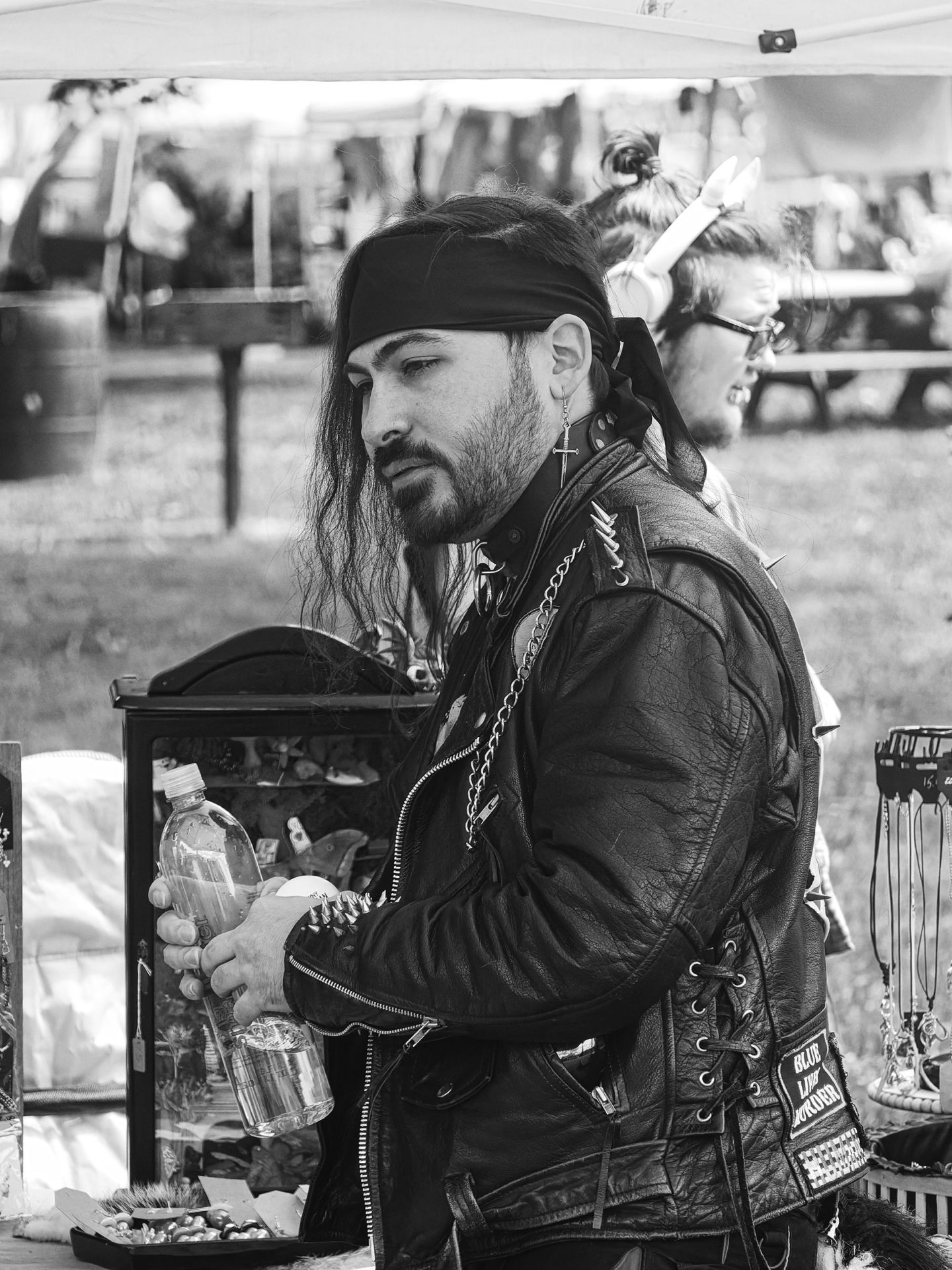 Black-and-white vertical shot of a man in a spiked leather jacket, black bandana, cross earring, and heavy chains, holding a plastic water bottle at what appears to be an outdoor vendor booth. A glass display case with small items sits to his left, jewelry hangs on a rack to the right, and a second person wearing glasses and headphones stands blurred in the background beneath a white tent canopy.