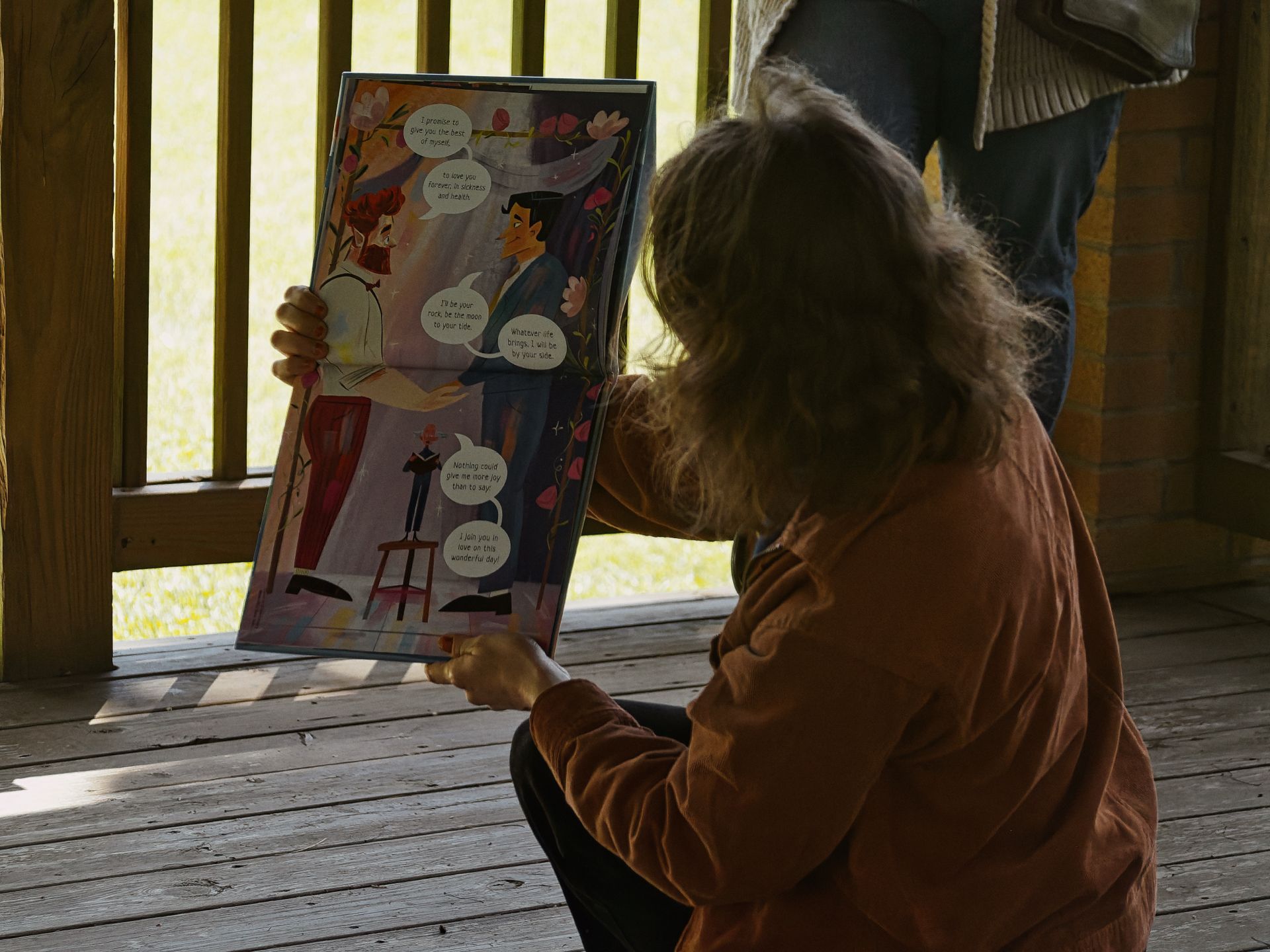 A child with wavy hair, wearing a rust-orange jacket, sits on a wooden porch deck holding open an illustrated book. The open spread shows a colorful wedding scene with two figures exchanging vows, speech bubbles reading "I promise to give you the best of myself," "To love you forever, in sickness and in health," "I'll be your rock, be the moon to your tide," "Whatever life brings, I will be by your side," "Nothing could give me more joy than to say," and "I join you in love on this wonderful day." A second person stands partially visible in the background, green grass visible through the porch railing.