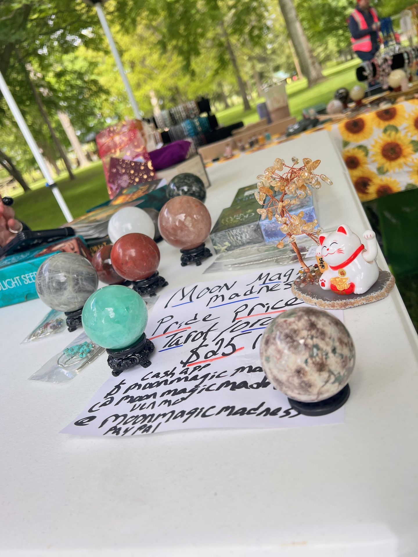 Outdoor market table covered in polished crystal spheres — including a pale aqua amazonite, a translucent clear quartz, a terracotta-red, and a speckled grey-green — each resting on small black carved stands. A handwritten sign in the foreground reads "Moon Magic Madness / Pride Prices / Tarot for... / $25 / Cash App @moonmagicma... / @moonmagicmadness / PayPal." To the right, a white ceramic maneki-neko lucky cat sits beside a small amber gemstone wire tree sculpture. A sunflower-print tablecloth, stacked books including one titled "LIGHT SEEDS," and a blurred green park setting fill the background.