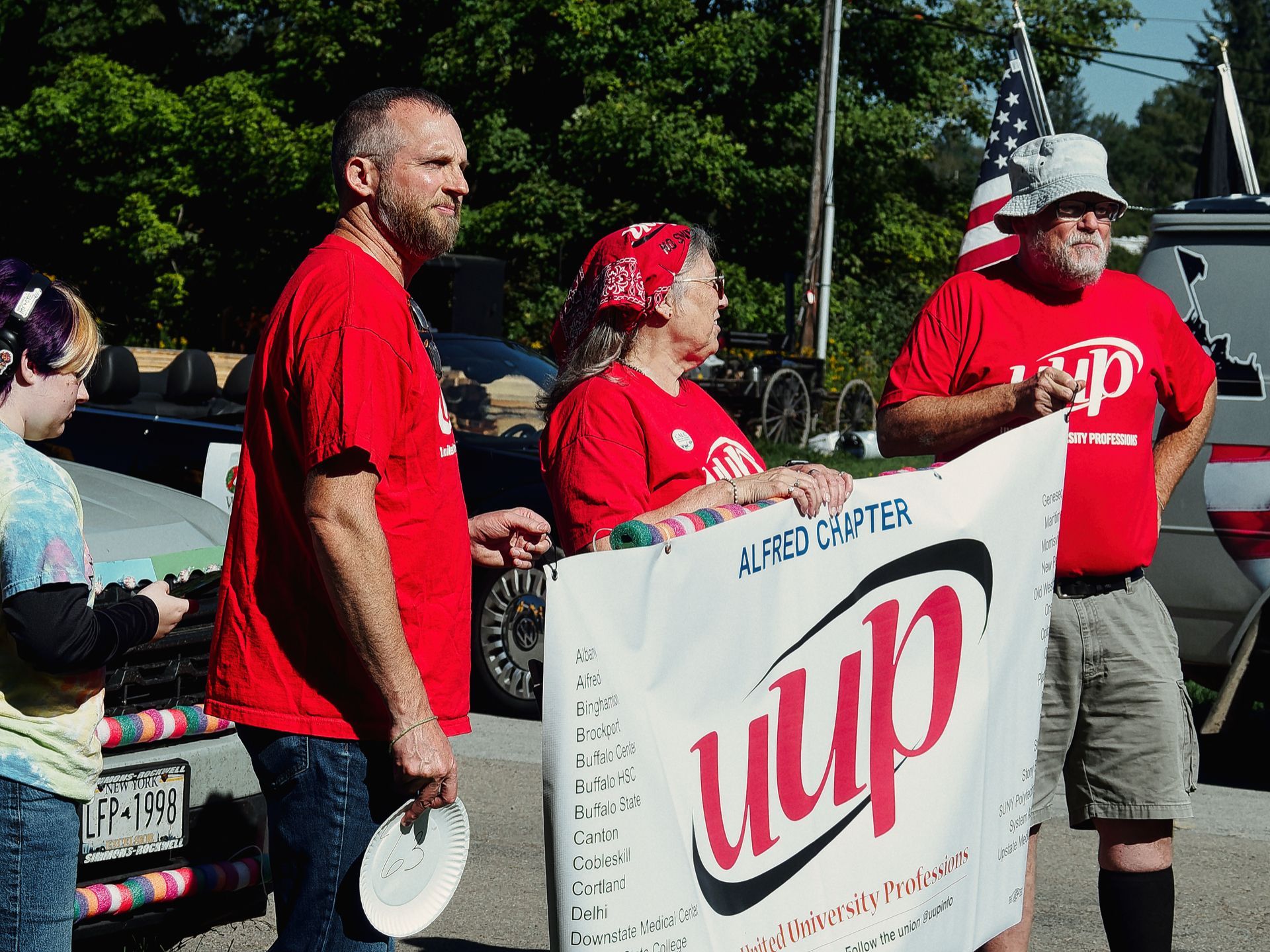 Three people in matching red UUP shirts hold a banner reading "Alfred Chapter / uup / United University Professions" at what appears to an outdoor event, with a partial American flag visible behind them. A fourth person in a tie-dye shirt stands at the left edge of the frame, and a New York license plate reading "LFP-1998" is visible on a car in the middle ground, with dense green trees filling the background.