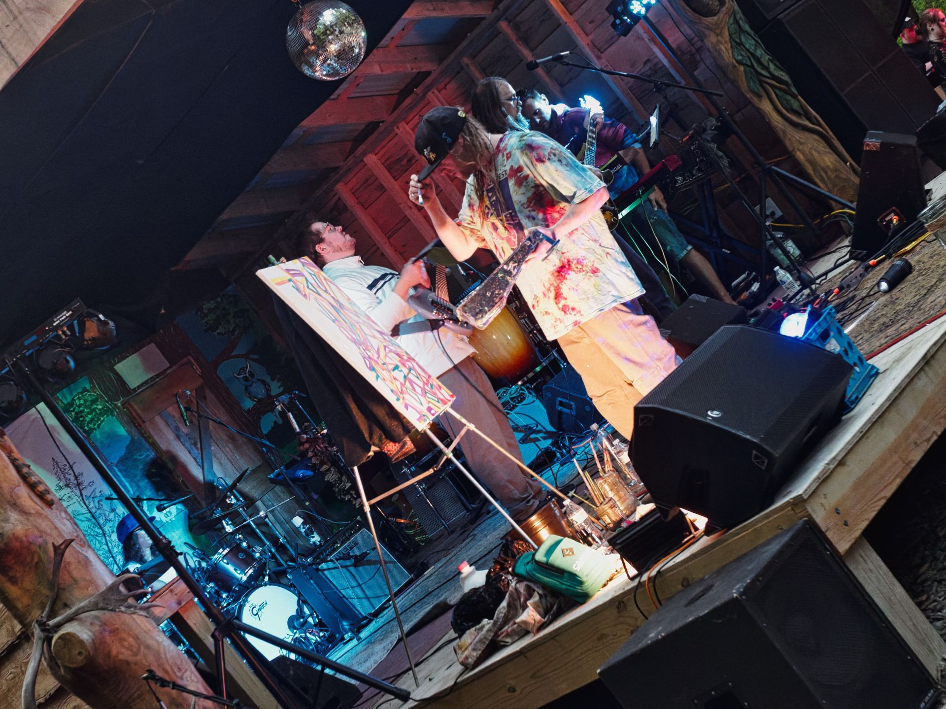 Rotated shot of a rustic indoor stage where a paint-splattered artist in a black cap holds a brush over a colorful abstract canvas on an easel, while a person beside them holds a microphone; a drum kit glows blue-green in the background left, a disco ball hangs from the ceiling above, and stage monitors flank the front edge of the platform.
