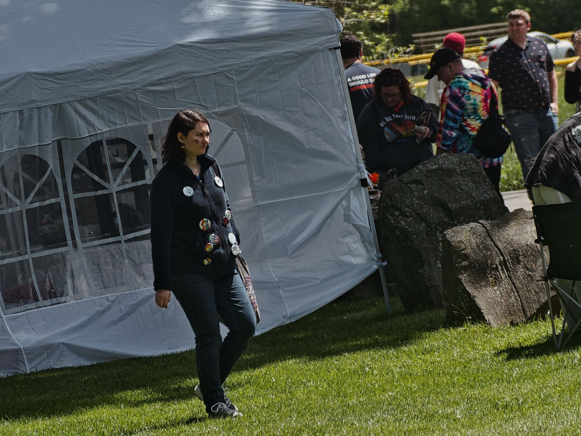 Woman in a black jacket covered in pinback buttons walking across a grass lawn in front of a white pop-up canopy tent, glancing to her left. Behind the tent, a small group mills around a table — one person in a tie-dye shirt, another in a black sweatshirt partially reading "A GOOD LIFE SHOULD B... HAPPENS" — with large boulders and a folding chair to the right.