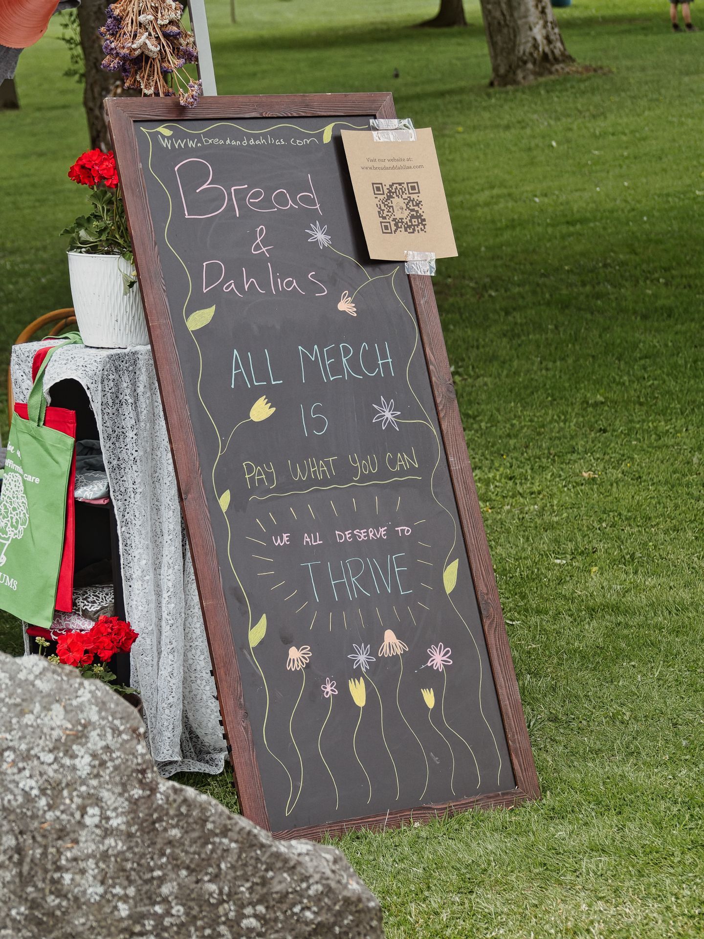 A tall chalkboard sign framed in dark wood, leaning against a vendor table at an outdoor market, reads "www.breadanddahlias.com / Bread & Dahlias / ALL MERCH IS PAY WHAT YOU CAN / we all deserve to THRIVE," decorated with hand-drawn flowers and leaves in pastel chalk. A kraft paper QR code card is taped to the upper right corner, and to the left, a table draped in lace holds red flowers and a white pot, with green and red tote bags hanging below.
