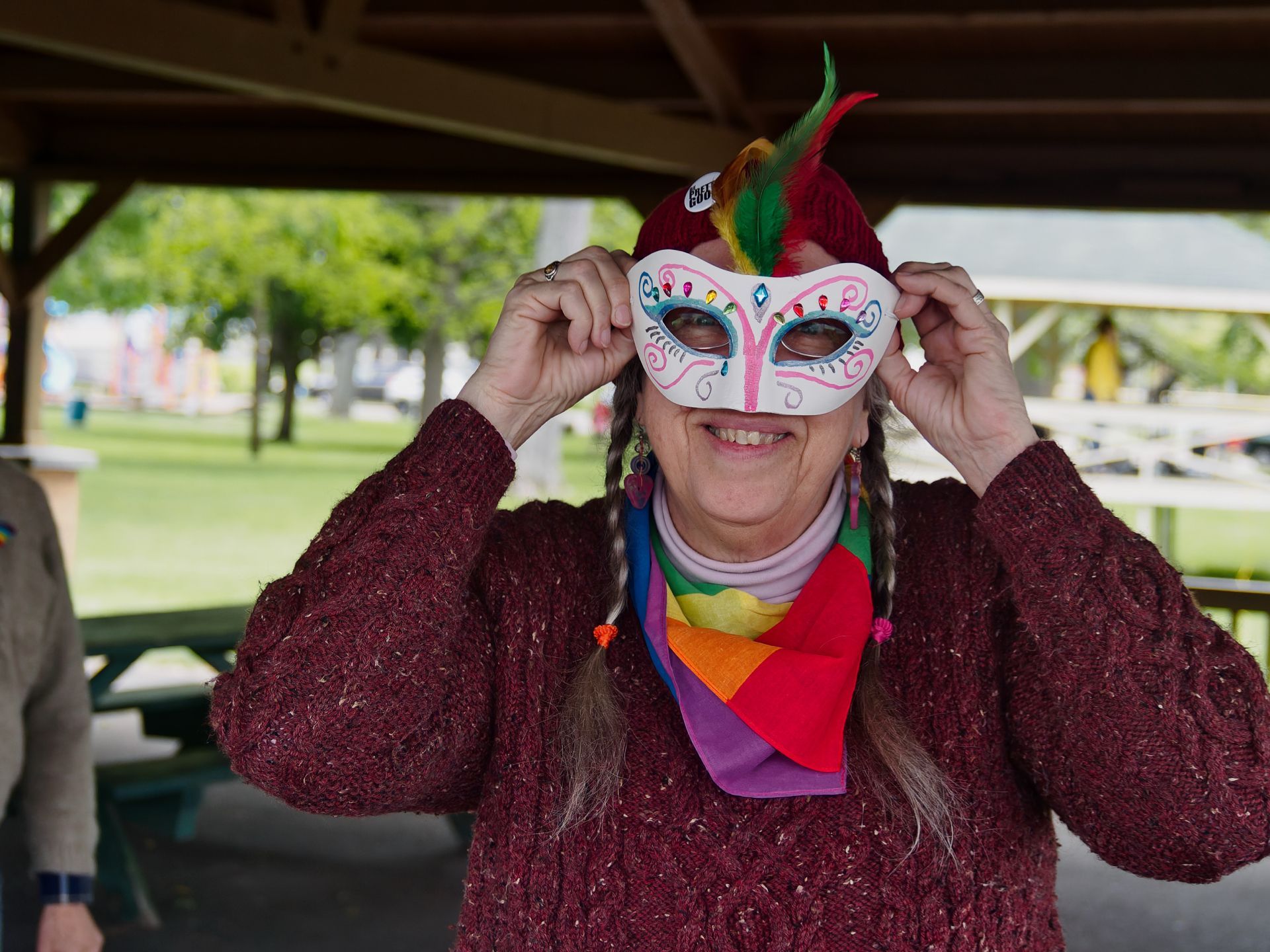 Woman in a burgundy knit sweater and rainbow scarf holding a white Mardi Gras-style masquerade mask up to her face, smiling broadly beneath it. The mask is hand-painted with blue and pink swirls and jewel details, topped with red, yellow, and green feathers. She stands under a wooden park pavilion, with green trees and lawn blurred softly in the background.