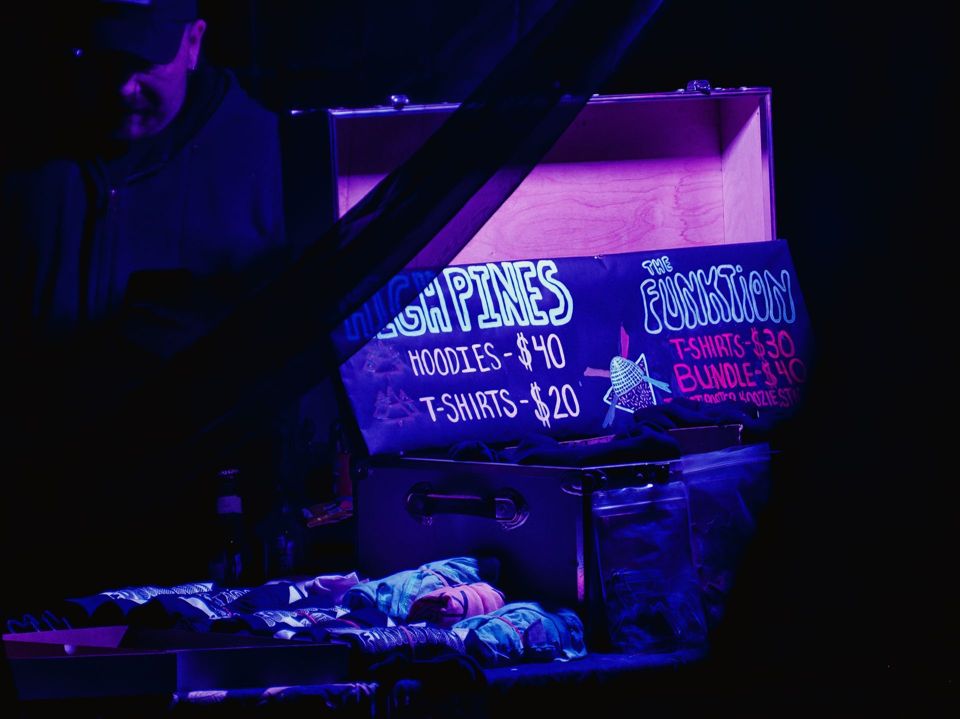 A merch table bathed in deep purple light, with an open wooden-lidded trunk painted black serving as a sign — left side reads "HIGH PINES / HOODIES - $40 / T-SHIRTS - $20," right side reads "THE FUNKTION / T-SHIRTS - $30 / BUNDLE - $40." Folded clothing is spread across the table in the foreground, and the silhouetted profile of a person in a cap is partially visible on the far left against the dark background.