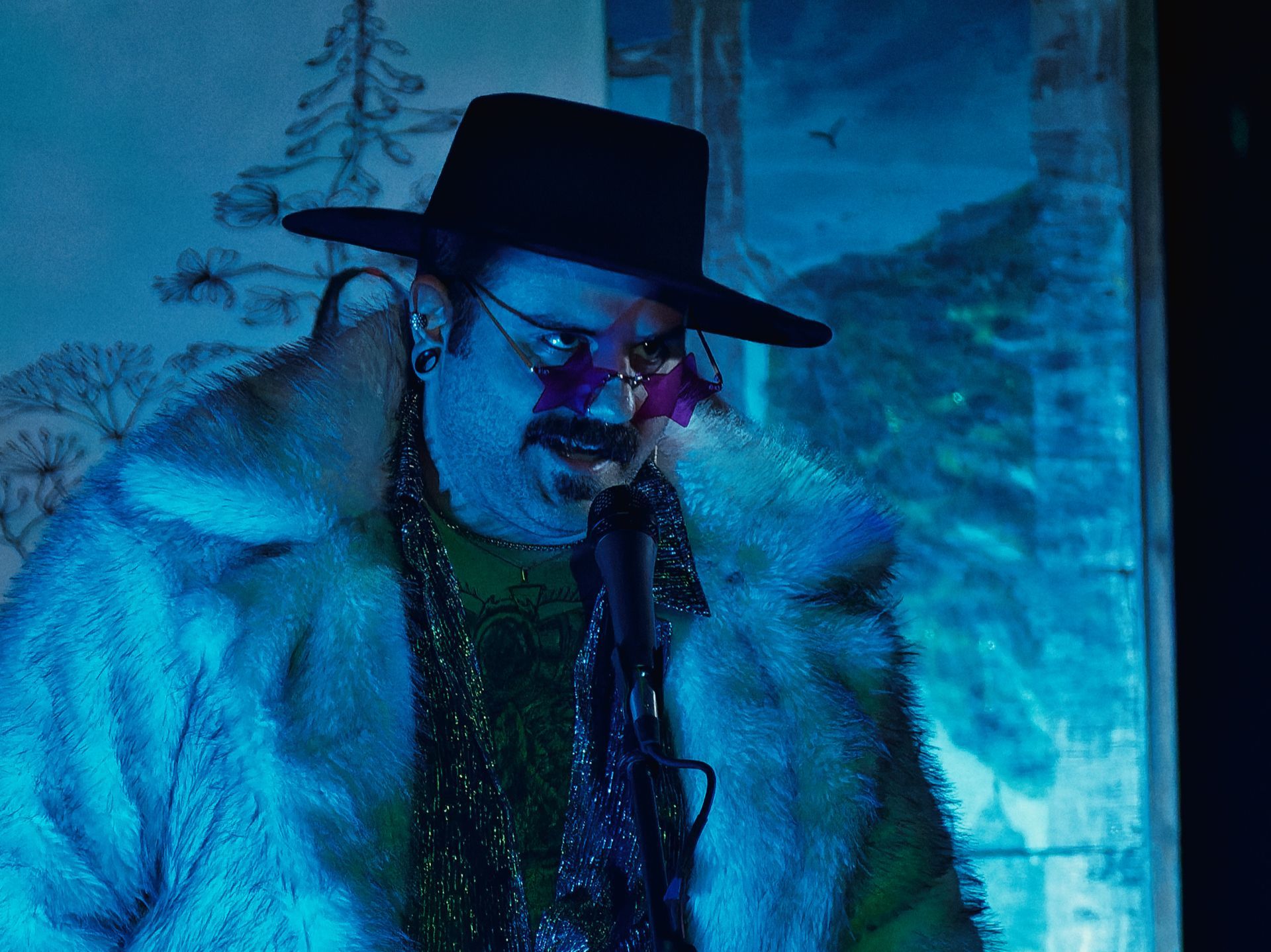 Person in a wide-brimmed black hat and star-shaped purple sunglasses, draped in a voluminous white fur coat over a green graphic top and shimmering silver vest, leaning toward a microphone stand under cool blue stage lighting, with a painted mural of trees and a landscape visible in the background.