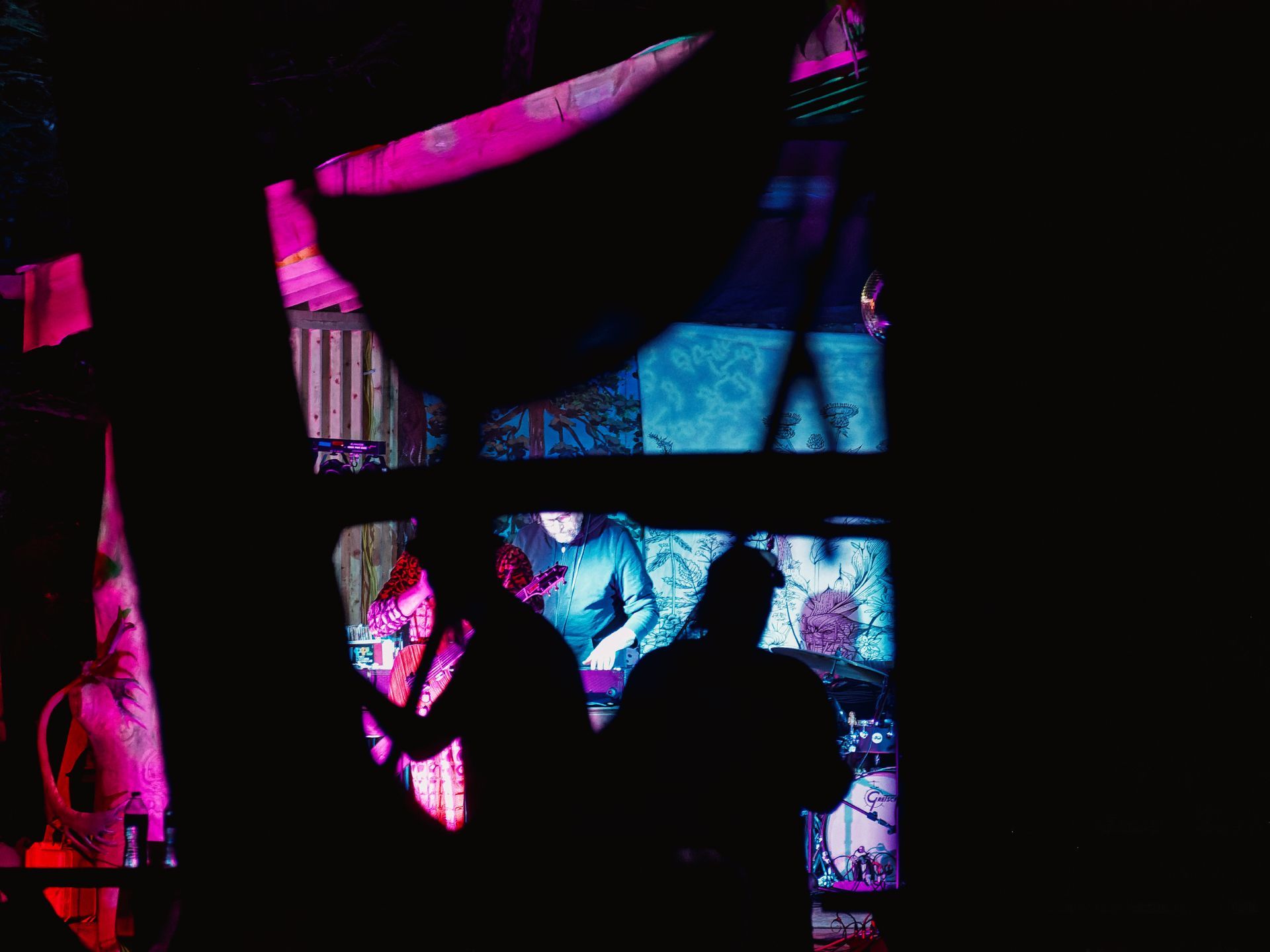 Darkened foreground silhouettes of audience members frame a fragmented, window-like view of a performer in a hoodie working equipment on a small stage, bathed in cool blue light against painted backdrop panels with illustrative designs, while deep magenta light bleeds in from the left where a second figure holds a guitar.