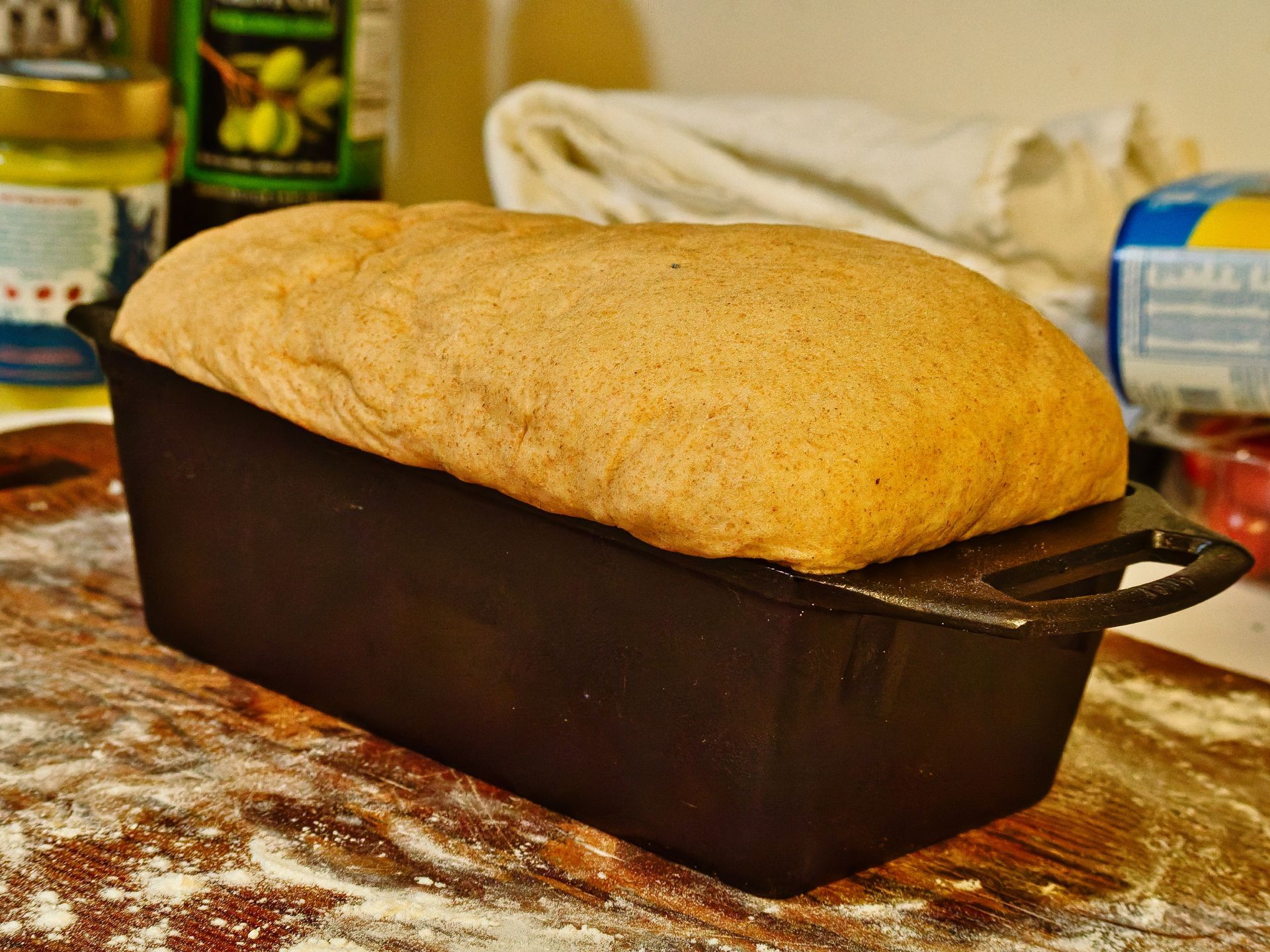 A risen bread loaf, golden-tan and dramatically domed, overflowing a black cast iron loaf pan on a flour-dusted wooden surface. A green olive oil bottle, a can, and a blue-labeled container sit blurred in the background.