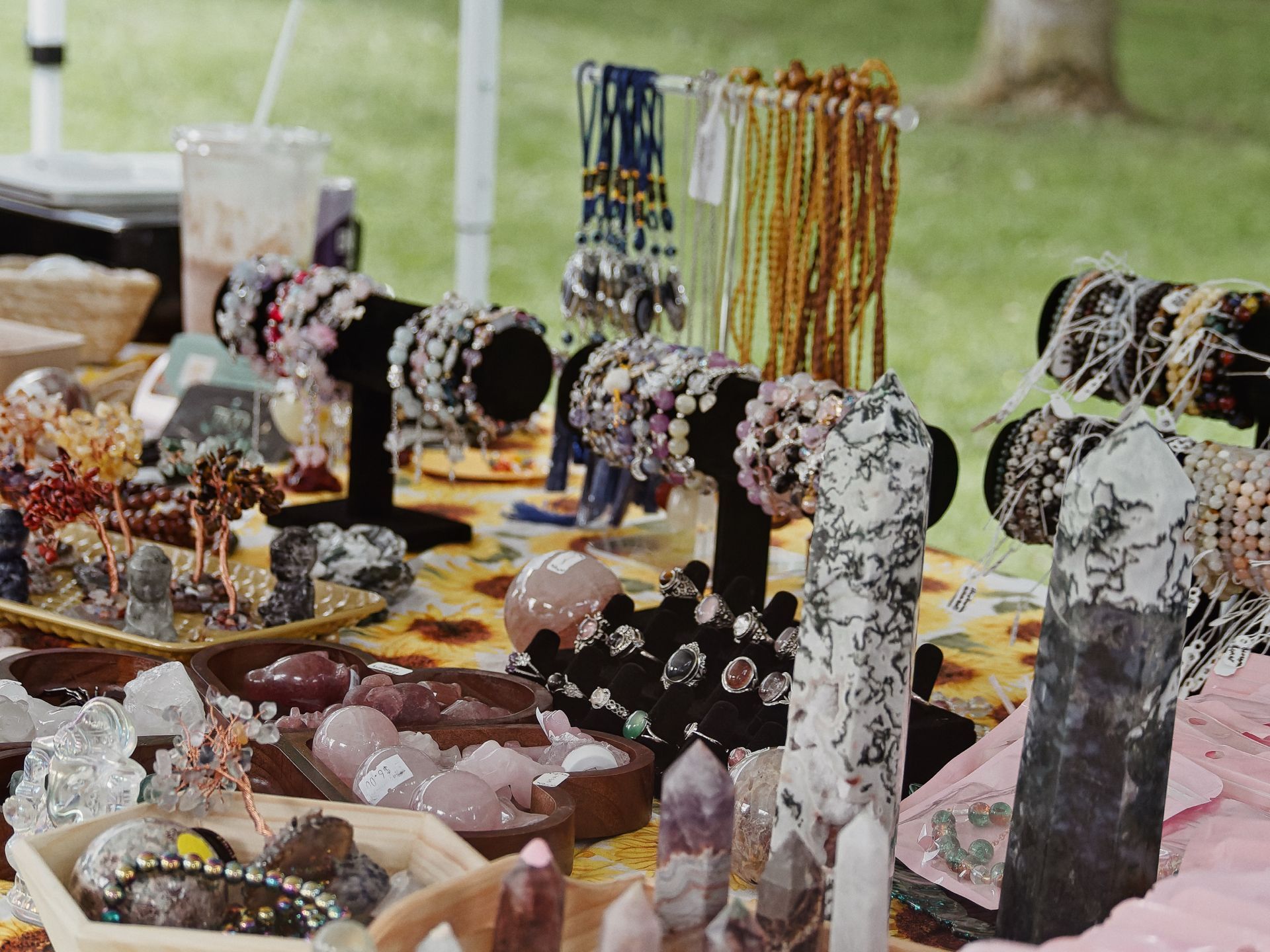 An outdoor market table densely covered in crystal and gemstone jewelry — polished rose quartz towers, amethyst points, and snowflake obsidian obelisks in the foreground, with wooden bowls of tumbled rose quartz stones, silver cocktail rings on display stands, and wire-wrapped crystal tree figurines to the left. Behind them, black velvet bracelet busts hold beaded and crystal-studded bracelets, while tall spindle racks hung with amber and navy beaded necklaces rise toward the back, blurred against a green grassy background.