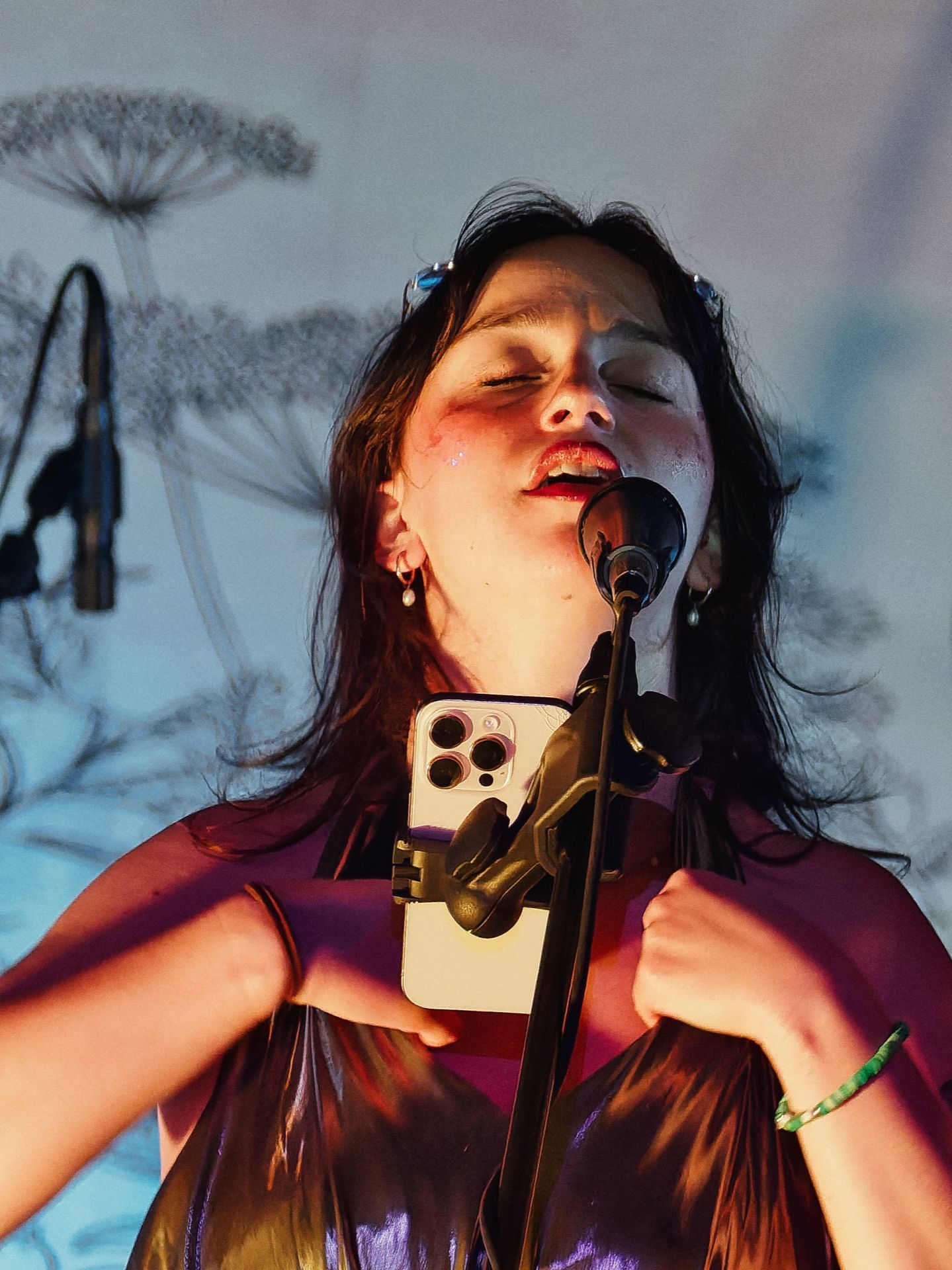 A dark-haired woman singing into a microphone with eyes closed and red lips slightly parted, visibly emotional under warm stage lighting, wearing a shimmery purple-black top and a green beaded bracelet, with an iPhone mounted on the mic stand in front of her chest. Behind her, a pale blue projection of silhouetted Queen Anne's lace plants fills the backdrop.