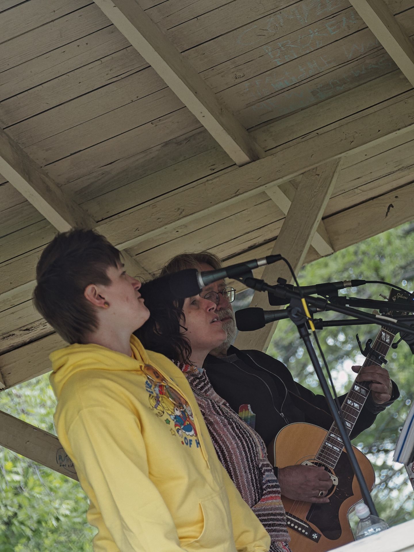 Two performers under a weathered wooden pavilion roof: a younger person in a yellow hoodie stands to the left, while an older musician with glasses and long dark hair plays an acoustic Gibson guitar and sings into a cluster of microphones on the right. Trees are visible and softly blurred in the background.