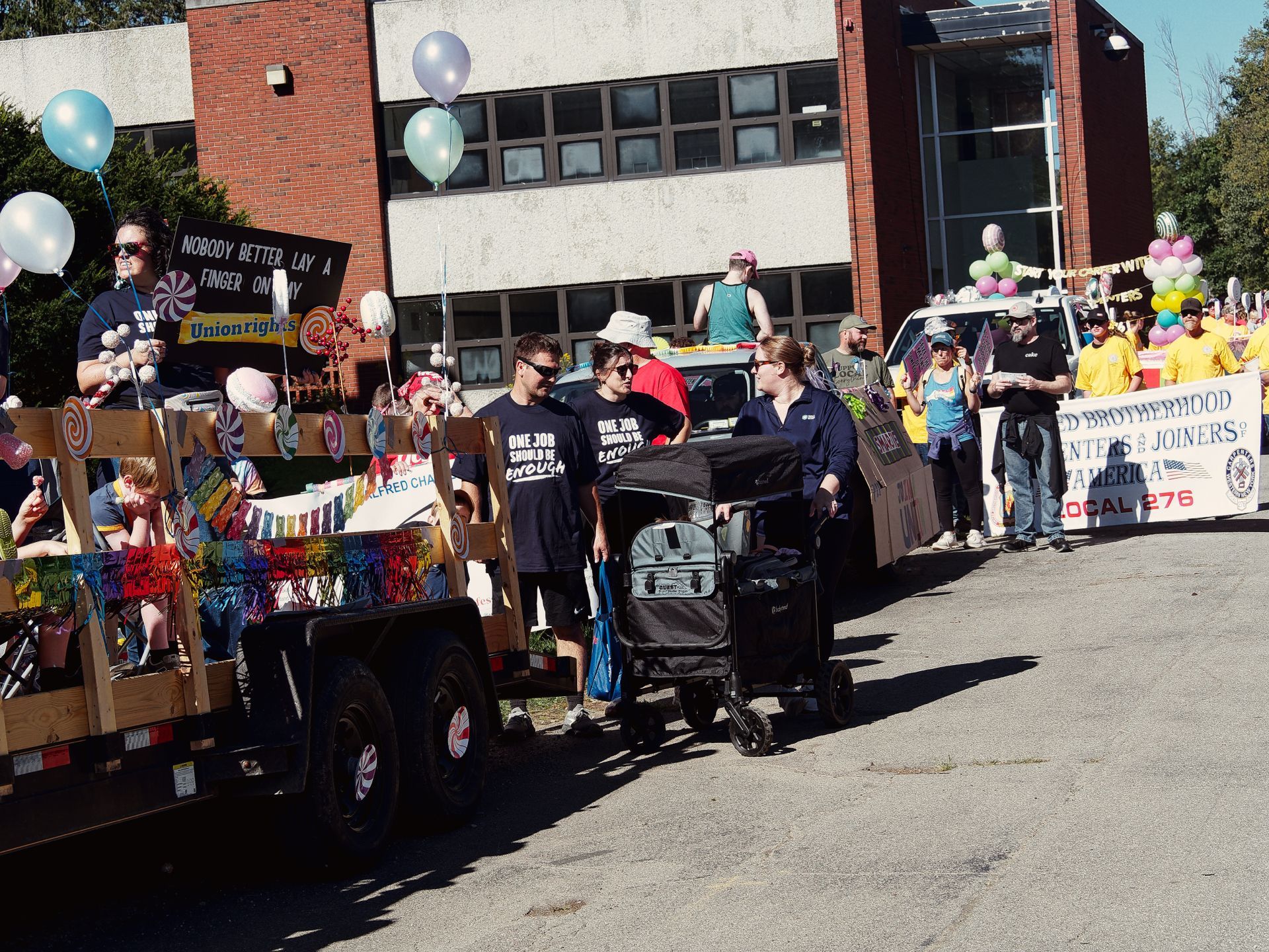 A Labor Day parade scene on a sunny street, with a candy-themed float on the left carrying children and decorated with lollipops and rainbow fringe, bearing a sign reading "Nobody Better Lay a Finger on My Union Rights." Two marchers in matching black shirts reading "One Job Should Be Enough" walk alongside a woman pushing a double stroller. To the right, a group in yellow shirts marches behind a banner reading "Brotherhood of Carpenters & Joiners of America Local 276," with colorful balloons overhead and a brick institutional building in the background.