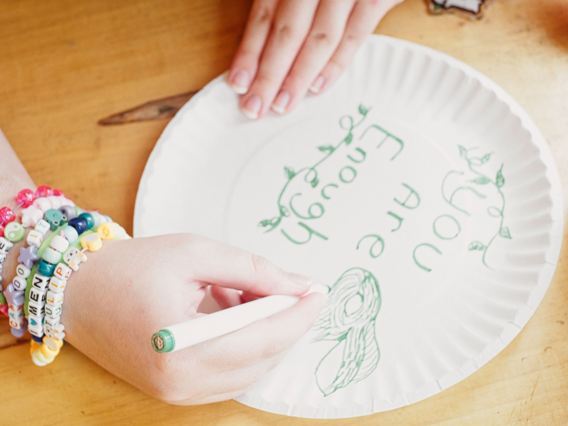 A hand adorned with layered beaded bracelets — including letter bracelets reading "I <3 MEN" and "TULIP" among others — uses a green marker to write "You Are Enough" on a white paper plate, decorated with small vine and leaf doodles and a sketch of a rabbit's face at the bottom. A second hand steadies the plate from the top, resting on a wooden surface.